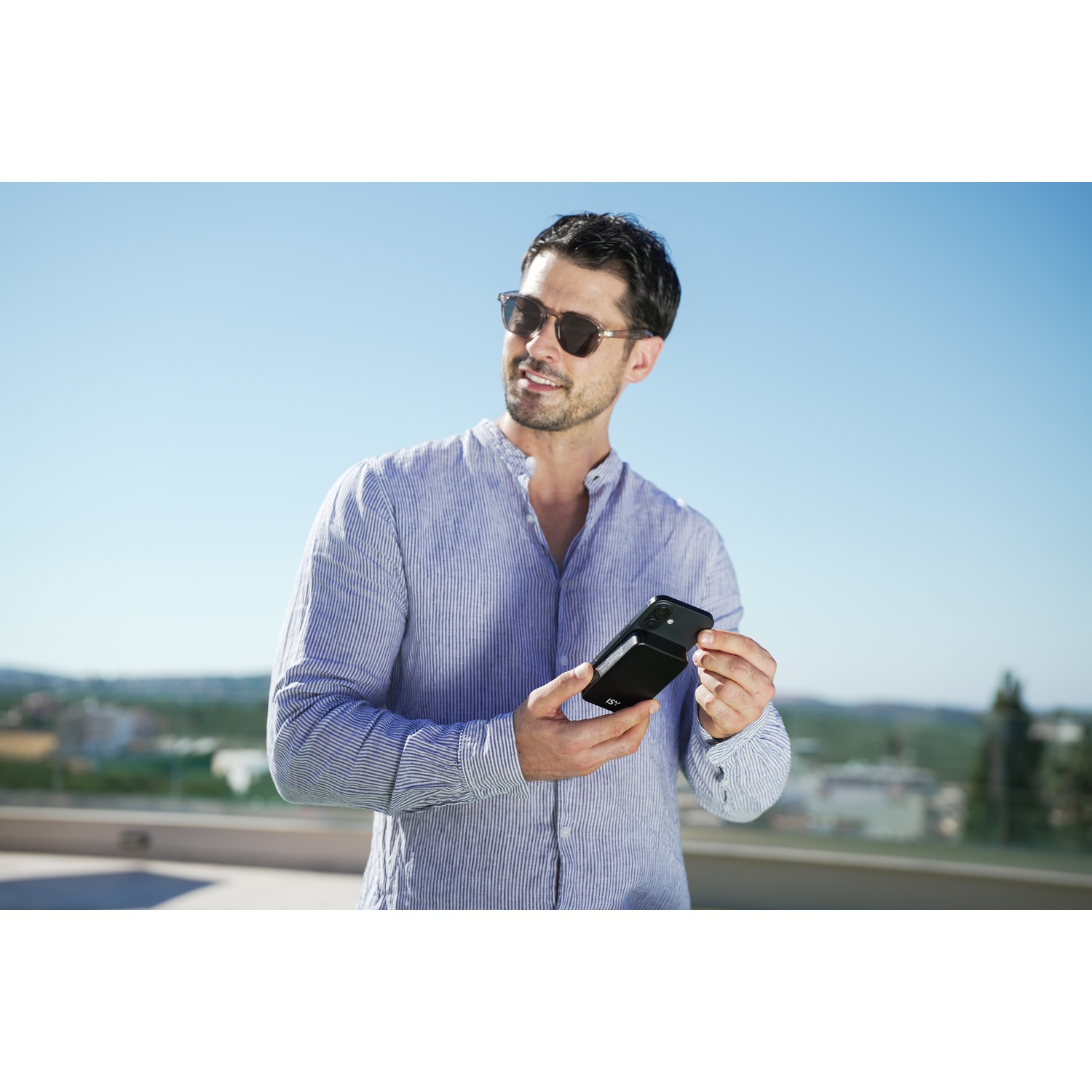 Un uomo con occhiali da sole e una camicia a righe blu tiene un telefono. Sorride. Lo sfondo è un cielo blu e degli edifici.