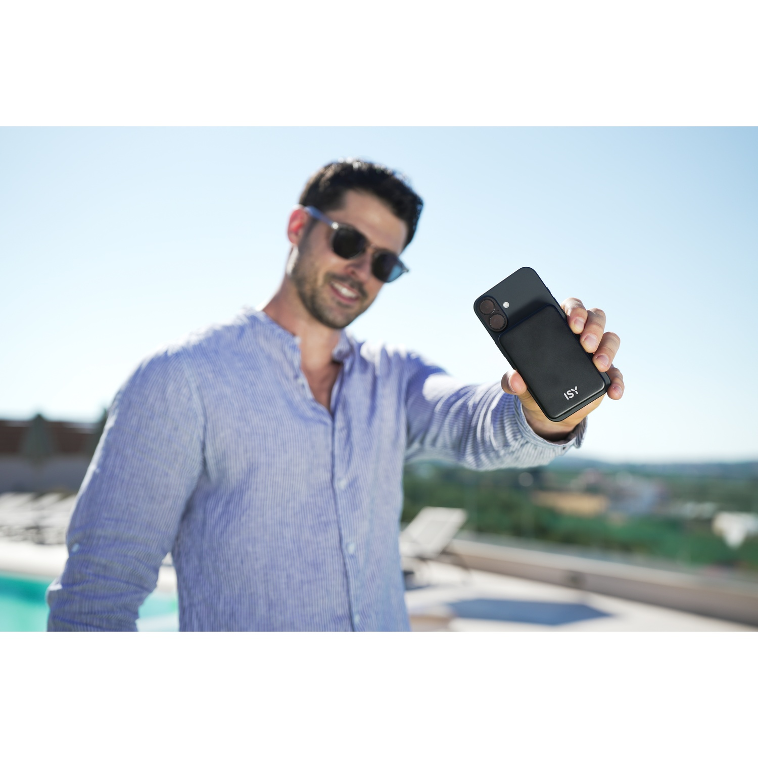 Un uomo con occhiali da sole tiene un telefono nero. Sorride. Cielo blu e sfondo sfocato con alberi visibili.