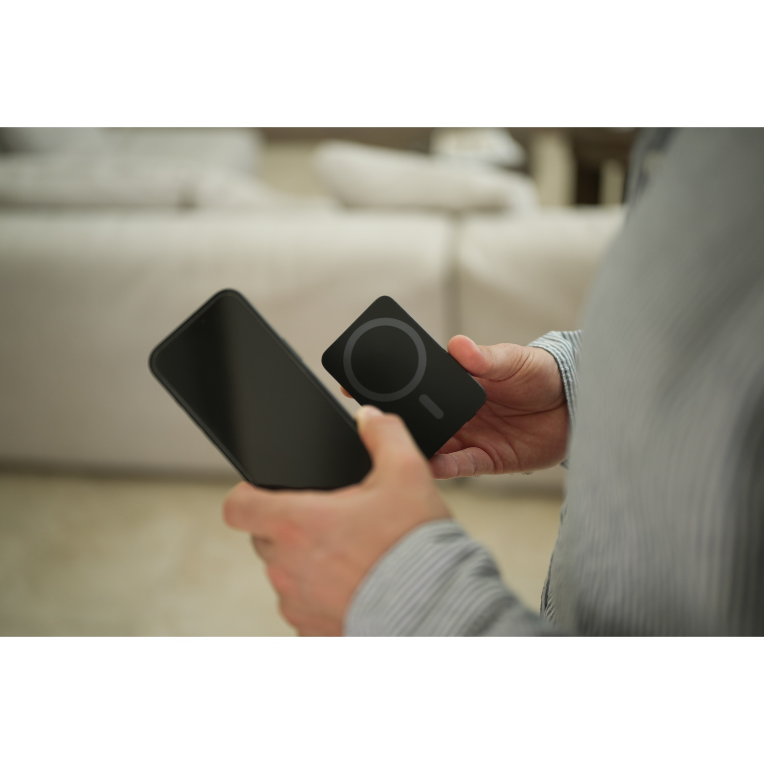 Una persona tiene in mano uno smartphone nero e un caricabatterie MagSafe nero. Sullo sfondo c'è un divano chiaro.