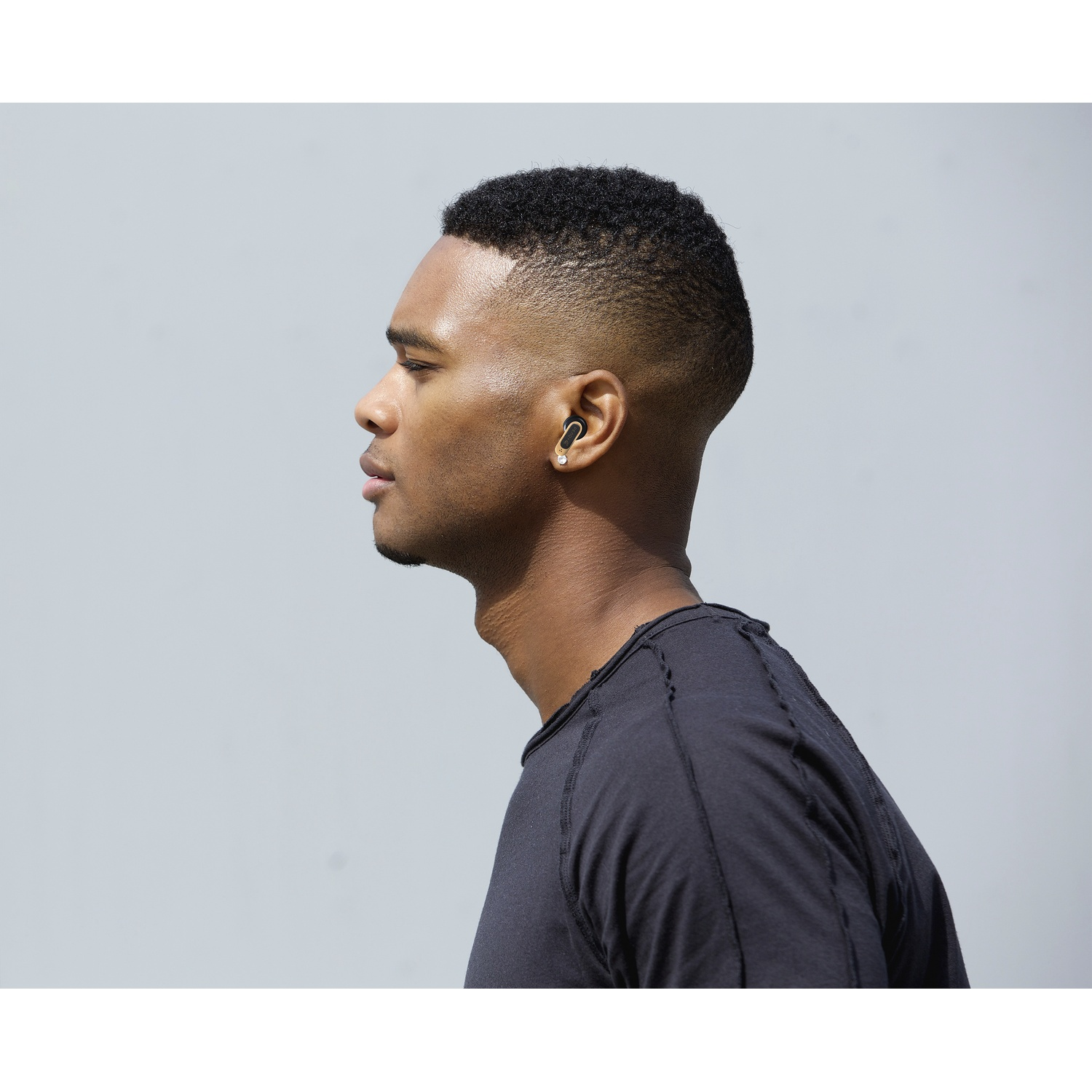 A man in a black shirt wearing a black earbud, looking to the left. Background is a solid, light grey color.