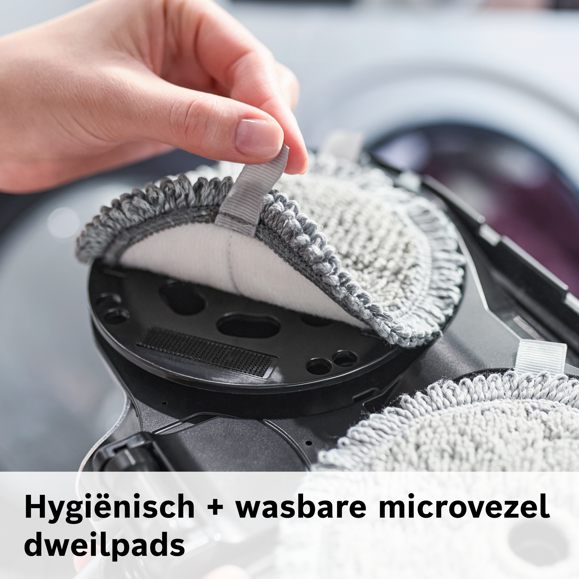 A hand lifts a microfiber pad from a black base. The pad is gray and white. The background is blurred.
