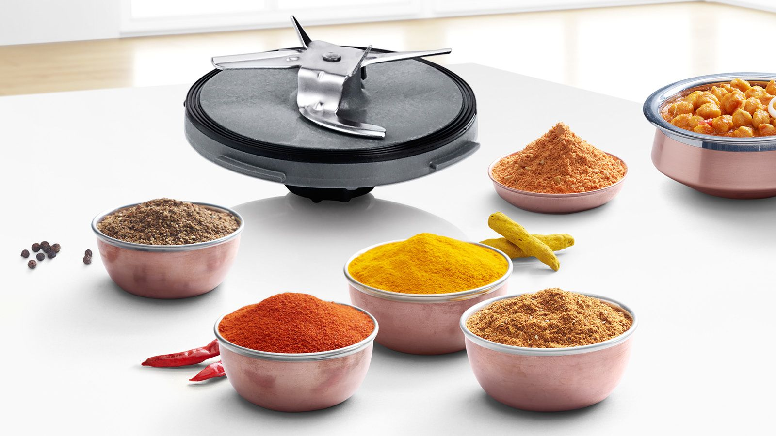 Een blenderblad staat boven kommen vol specerijen. Specerijen zijn onder meer rode, gele en bruine poeders, plus kurkuma en zwarte peperkorrels.