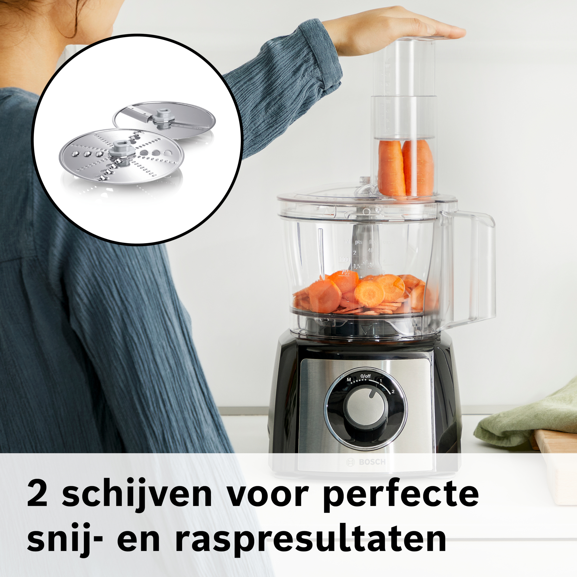 Een keukenmachine met wortels die worden gesneden. Twee messen worden in een cirkel getoond. Een hand plaatst wortels erin.
