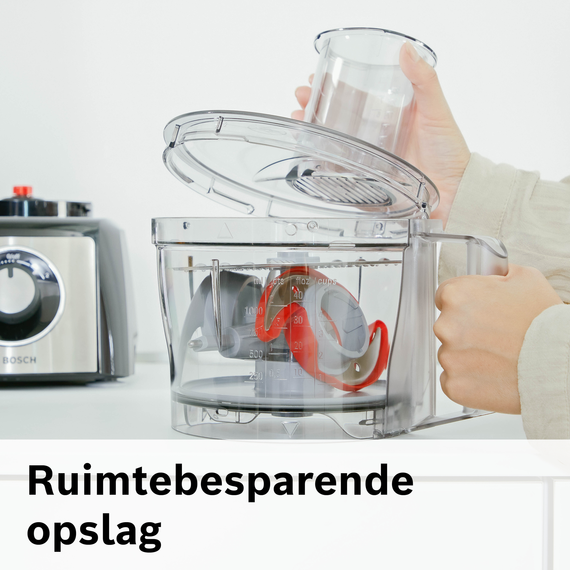 Een persoon opent een transparante keukenmachine met rode en grijze onderdelen en een ander keukenapparaat op de achtergrond.