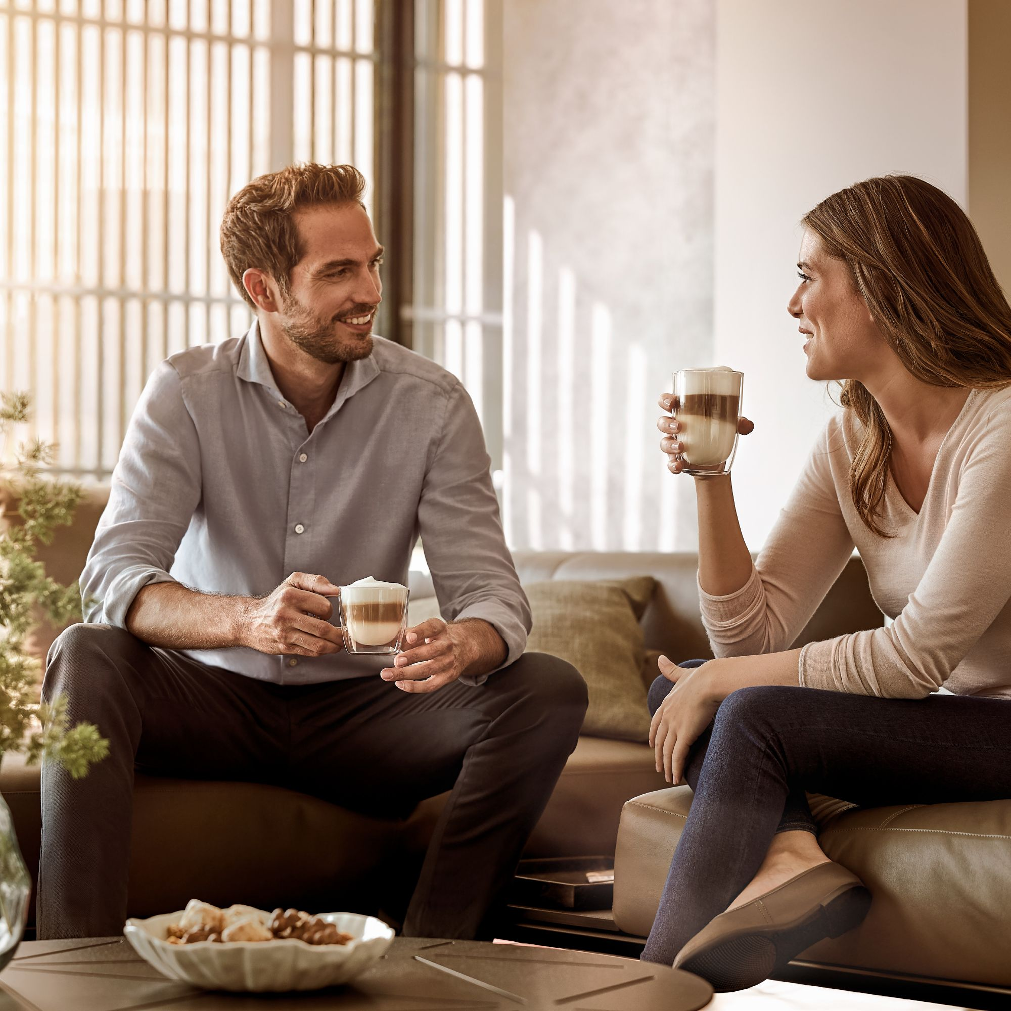 Een man en een vrouw zitten, koffie vasthoudend. Ze praten in een goed verlichte kamer.