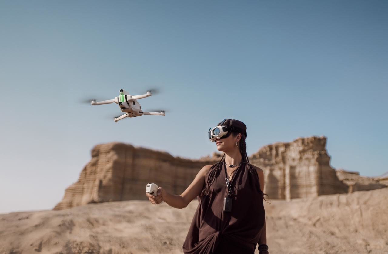 Una donna con occhiali VR controlla un drone con un telecomando. Il drone è sospeso sopra un paesaggio desertico sotto un cielo blu.