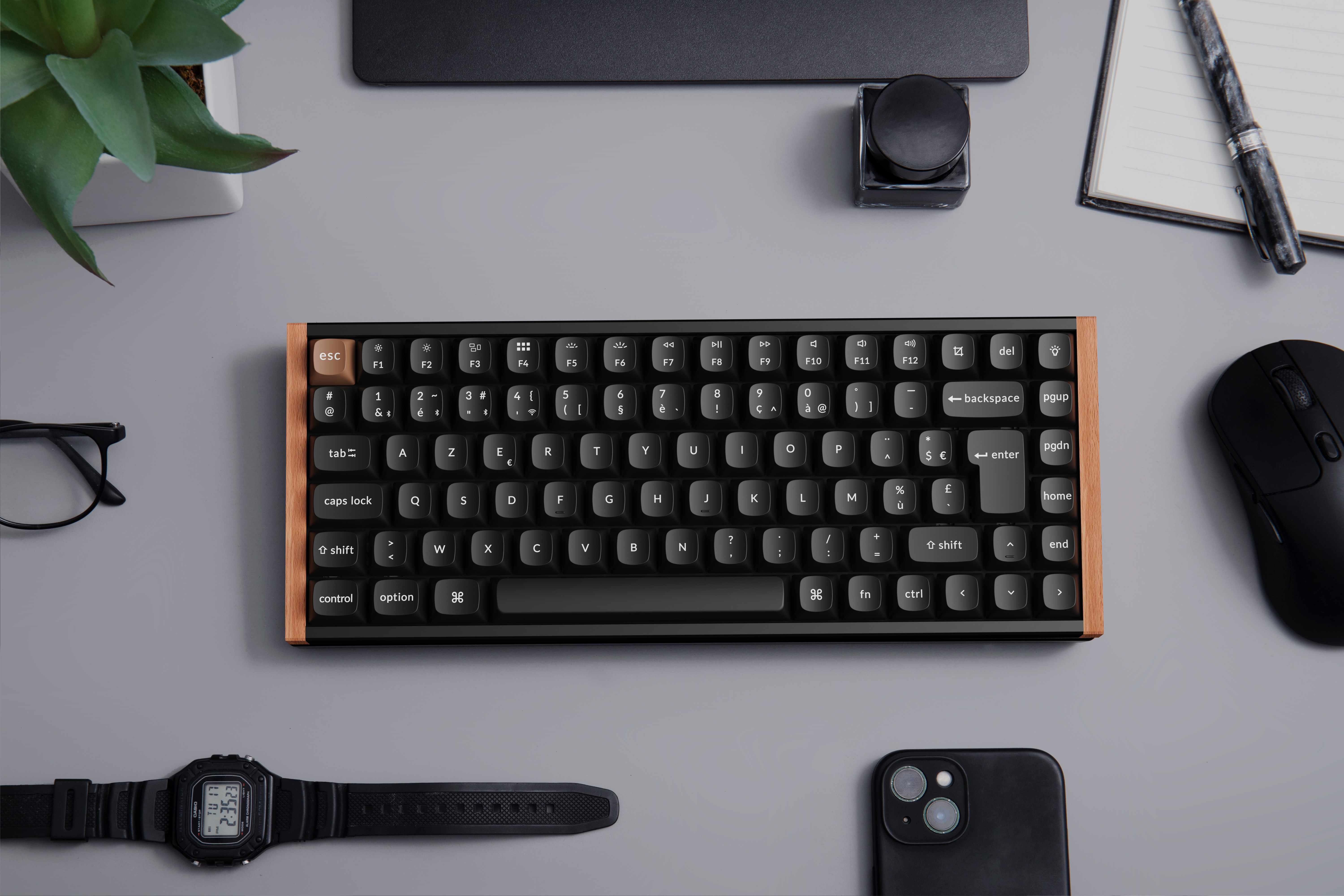 Draufsicht auf Tastatur, Uhr, Telefon, Brille, Maus, Notizblock, Tinte und Pflanze auf grauem Untergrund.