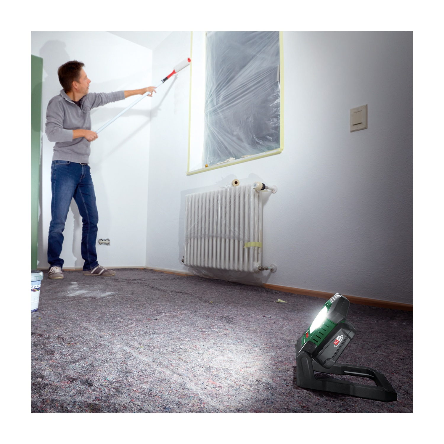 Una persona dipinge un muro con un rullo. Una lampada illumina la stanza. La finestra è coperta di plastica. Il pavimento è coperto.