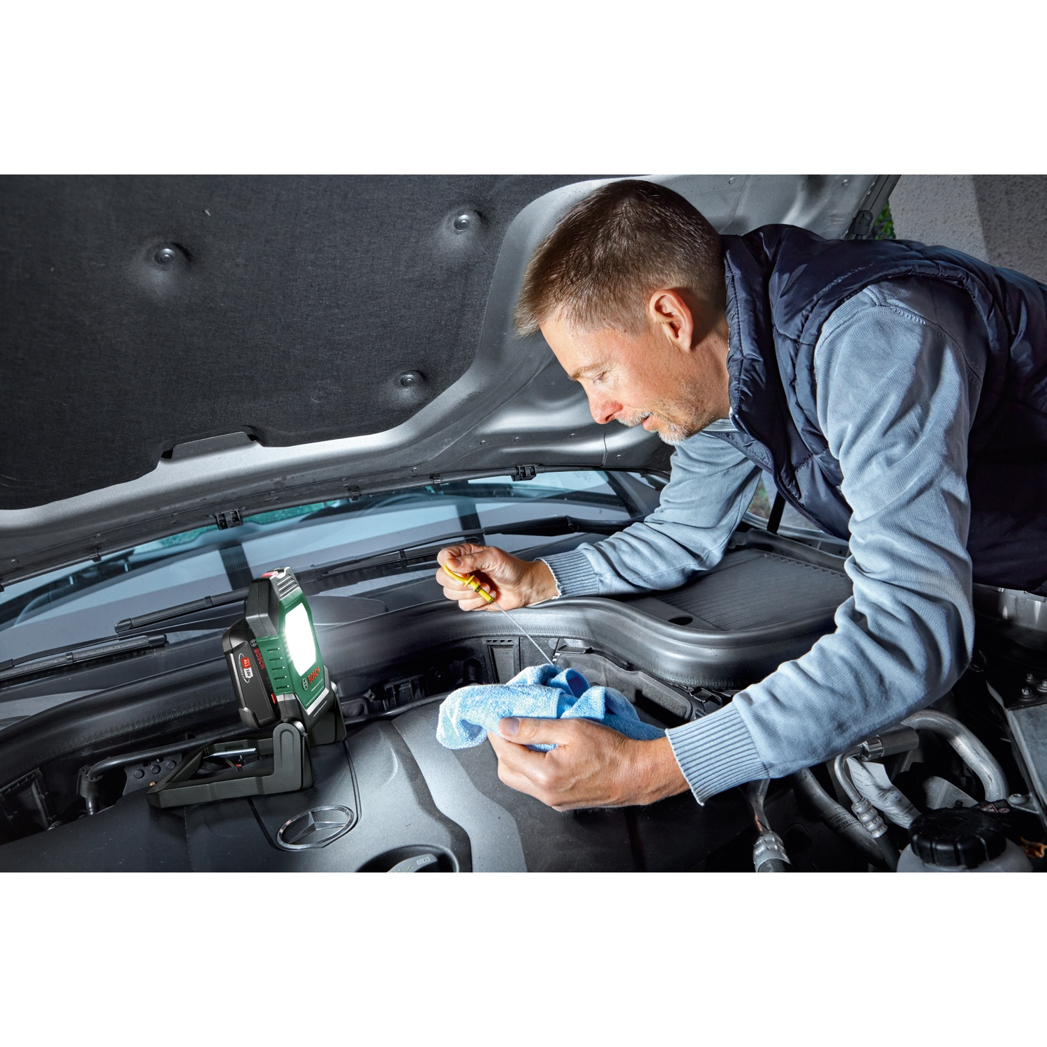 Uomo che lavora sul motore dell'auto, con panno blu. Luce da lavoro verde sul motore. Cofano aperto.