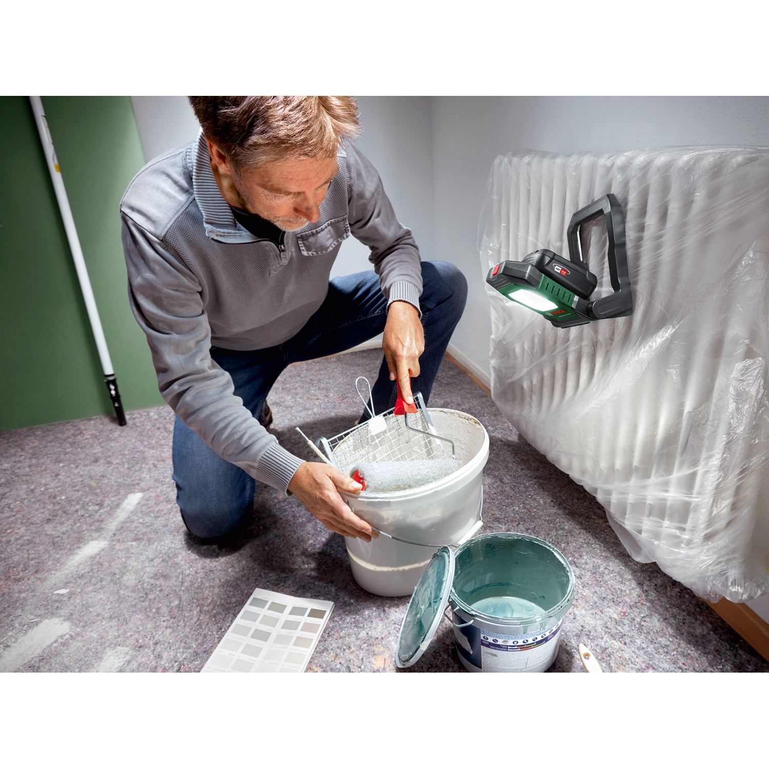 Un uomo si inginocchia preparandosi a dipingere. Secchi di vernice, rullo e campioni di colore sul pavimento. Una lampada da lavoro su un radiatore.