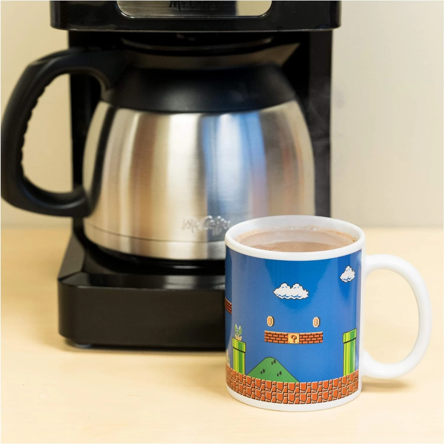 Cafetera con una taza de Mario. La taza tiene un fondo azul con un paisaje pixelado. Café en la taza.