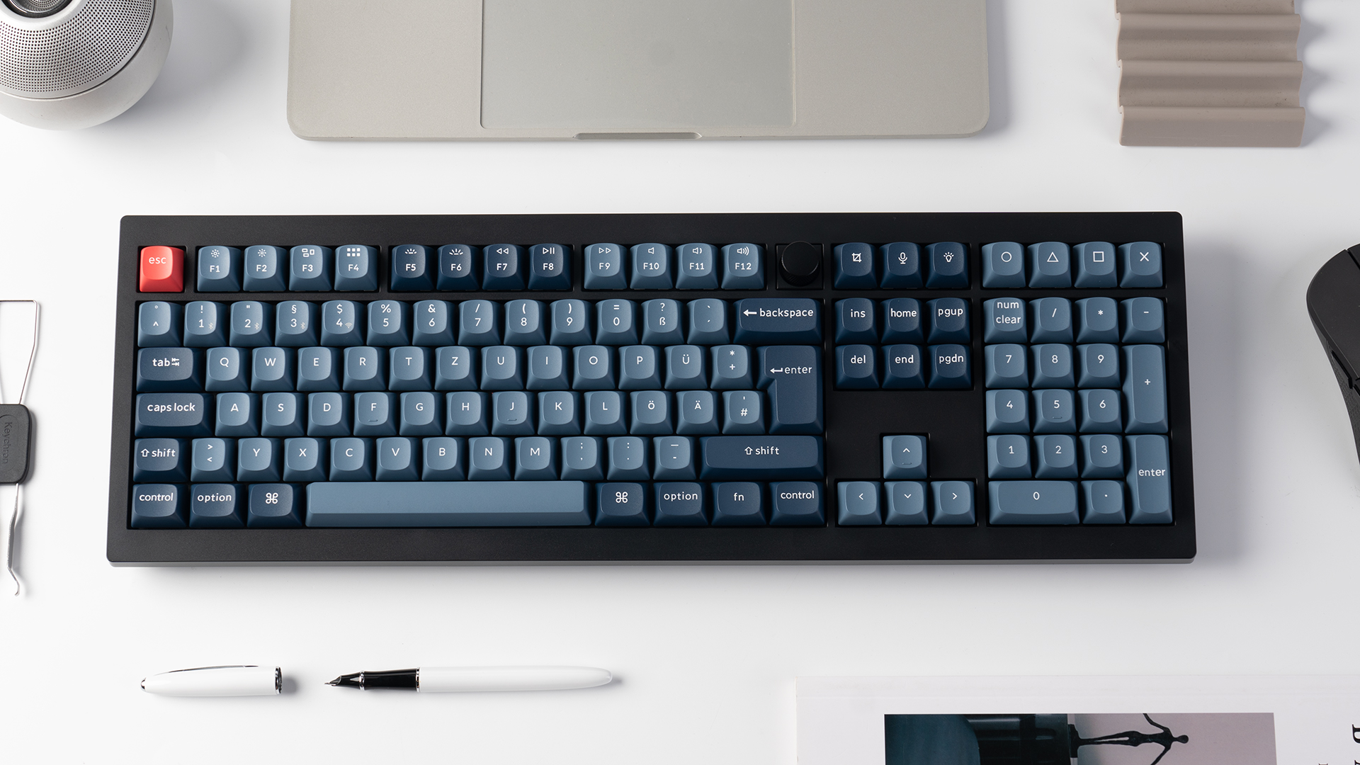 Eine schwarze Tastatur mit blauen Tasten, eine rote Escape-Taste. Weitere Artikel: Maus, Stifte und ein Laptop. Alles auf weißer Oberfläche.