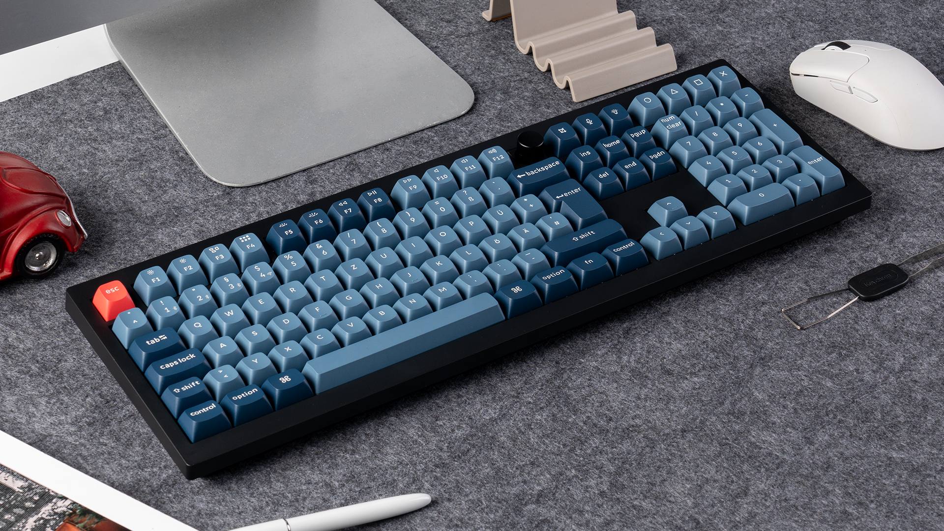 Eine schwarze Tastatur mit blauen Tasten, eine rote Escape-Taste, eine weiße Maus und ein rotes Auto auf grauem Untergrund.