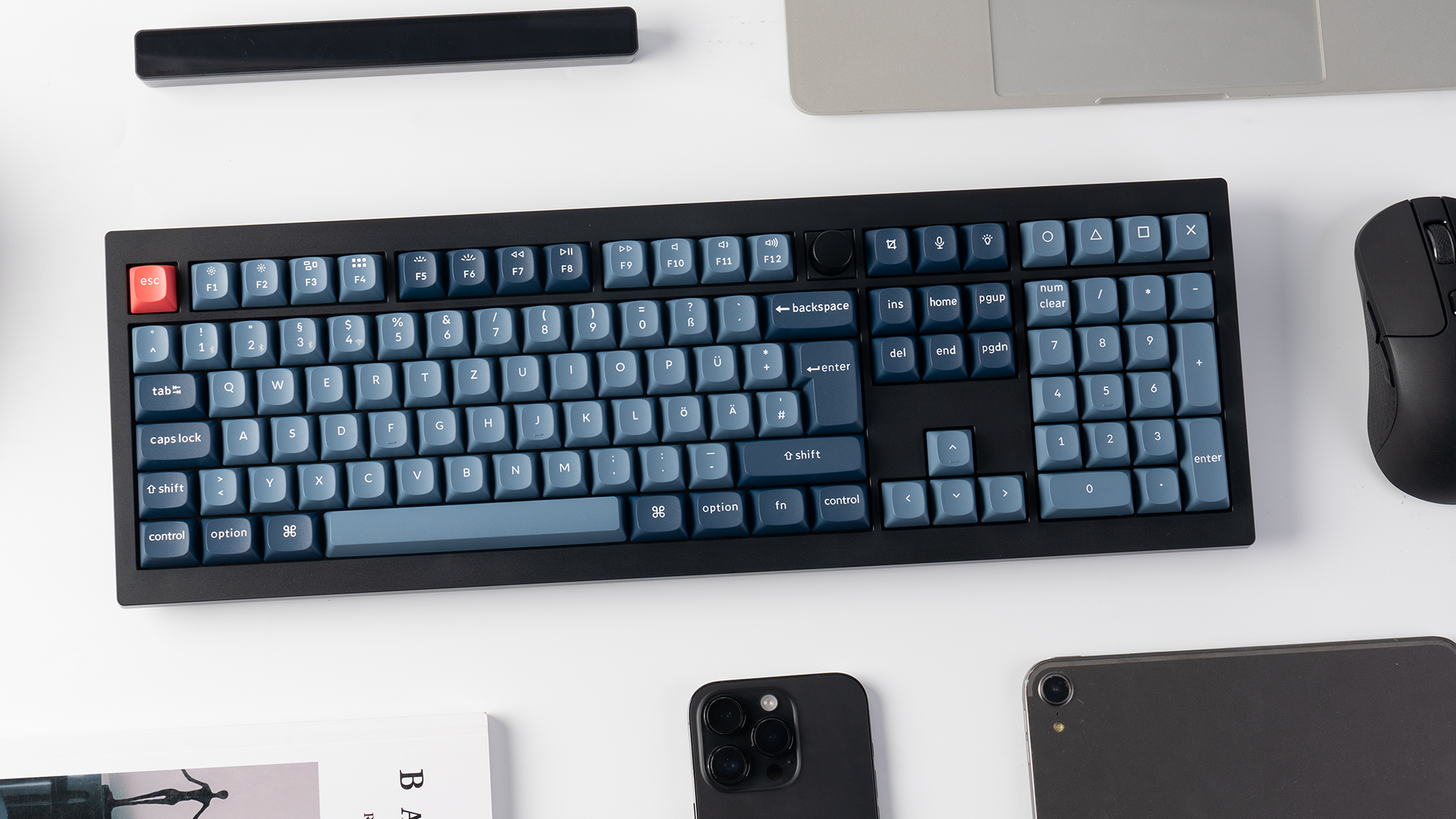 Eine schwarze Tastatur mit blauen Tasten, Maus, Telefon und Tablet auf weißer Oberfläche. Ein Buch und ein schwarzer Balken sind ebenfalls vorhanden.