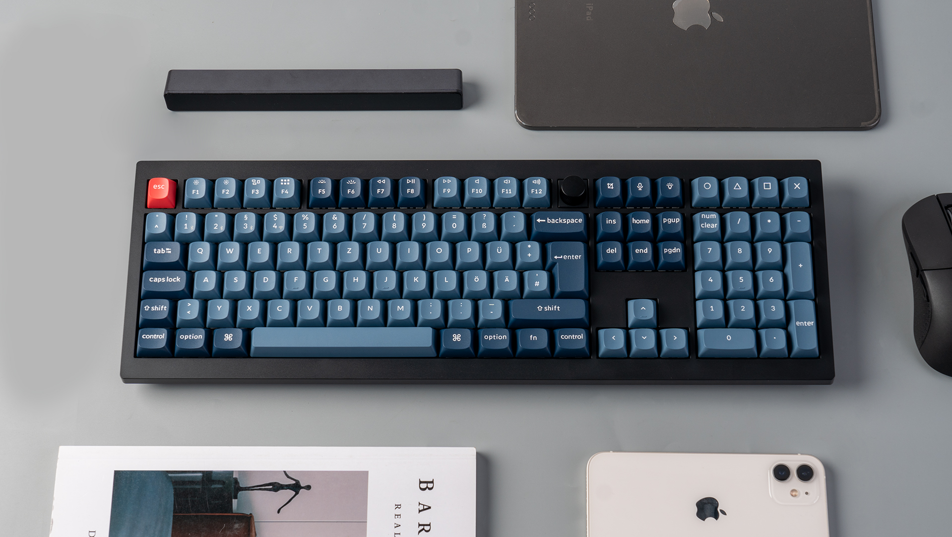 Eine schwarze Tastatur mit blauen Tasten, eine Maus, ein Tablet und ein Telefon liegen auf einer grauen Oberfläche. Ein Magazin ist unten.