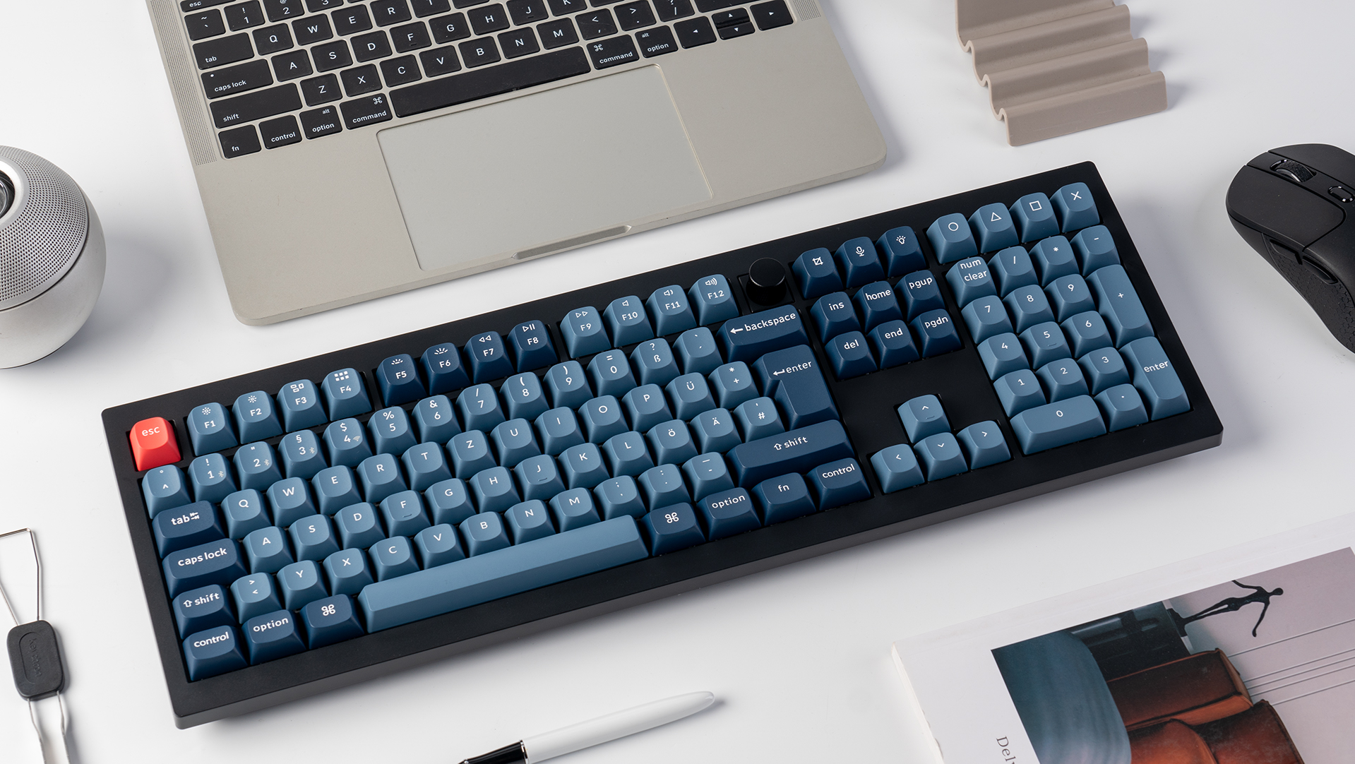 Eine Tastatur mit blauen Tasten und schwarzem Rahmen, ein Laptop, eine Maus und andere Gegenstände liegen auf einer weißen Oberfläche.