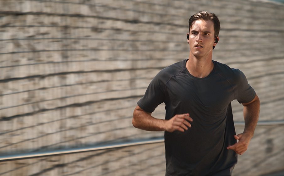 Mężczyzna biegnie w ciemnoszarej koszulce i słuchawkach. Tło to rozmyta drewniana ściana.