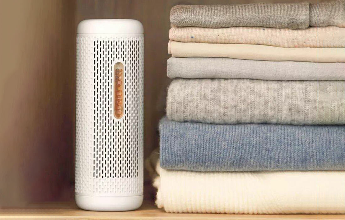 Biały oczyszczacz powietrza obok stosu złożonych swetrów w różnych kolorach, w tym szarym, beżowym i niebieskim, na półce.