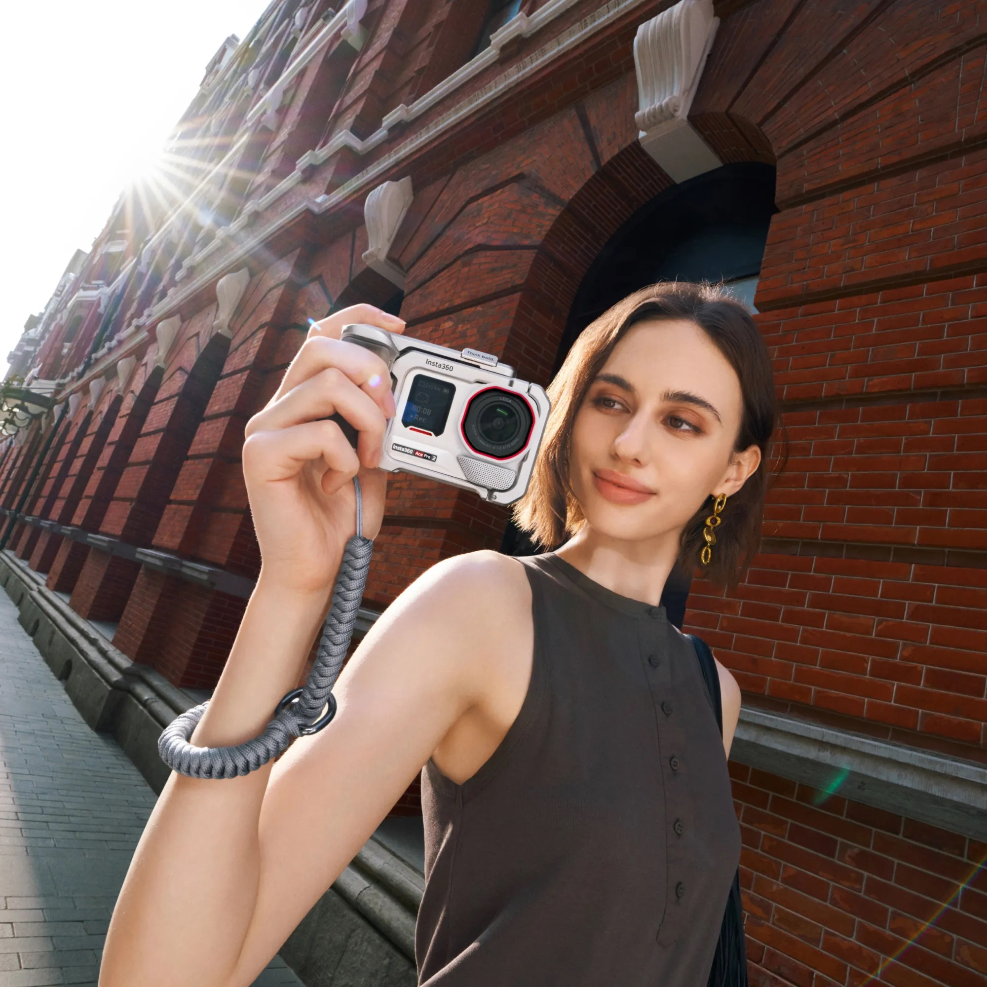 Kadın kamera tutuyor, selfie çekiyor. Arka planda kırmızı tuğla bina. Güneş parlıyor.