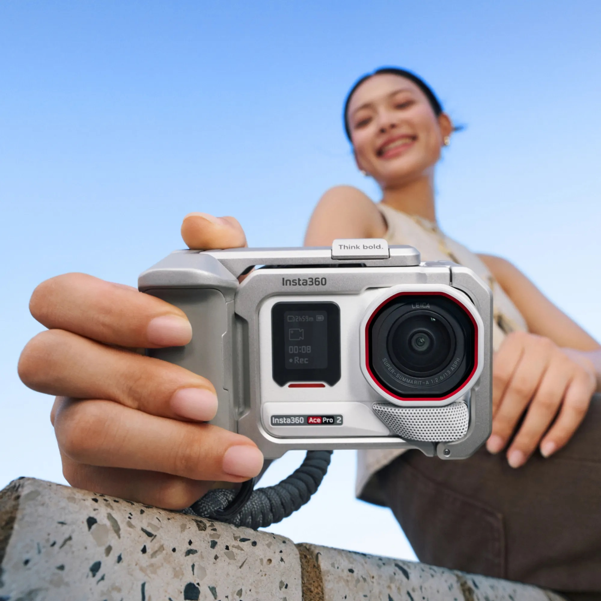 Bir kadın Insta360 Ace Pro 2 kamera tutuyor. Kameranın siyah bir lensi, kırmızı detayları var ve kayıt yapıyor. Arka planda mavi gökyüzü.