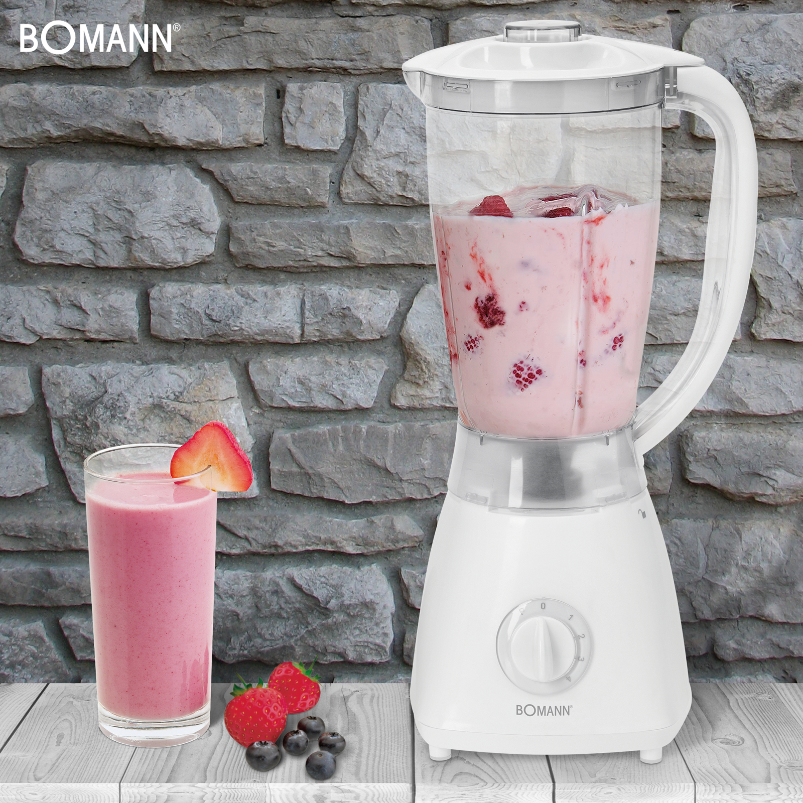 Ein weißer Mixer mit rosa Smoothie, ein Glas Smoothie, Erdbeeren und Blaubeeren stehen auf einem Holztisch.