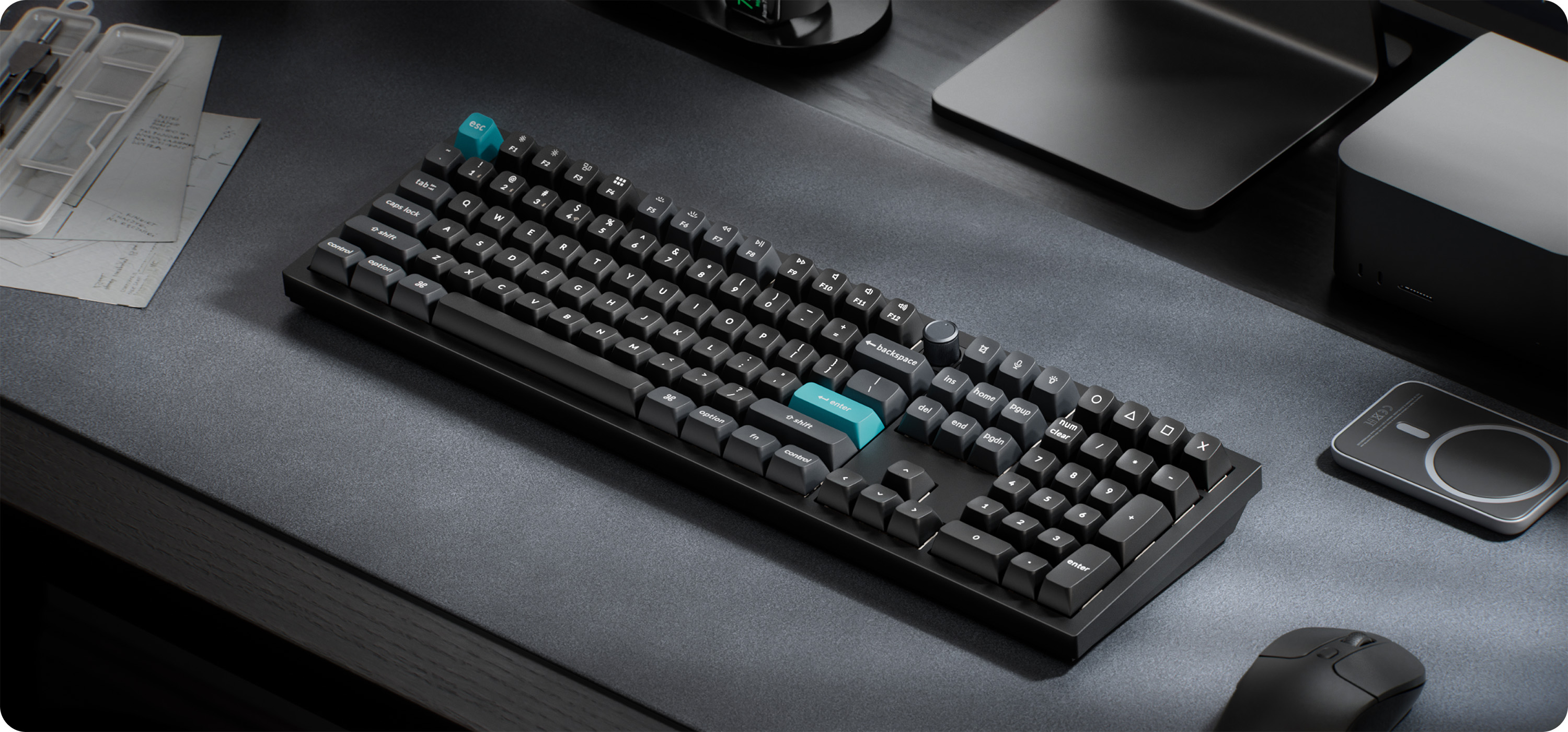 Schwarze Tastatur auf grauem Schreibtisch mit Maus, Laptop und Steuerungsknopf. Blaue Tasten sichtbar.
