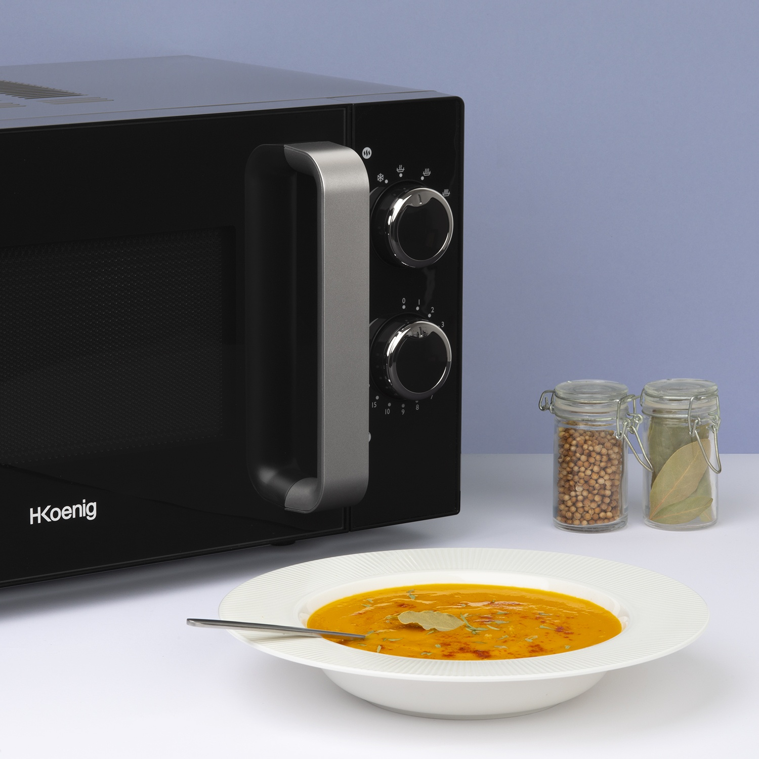 Un forno a microonde nero accanto a una ciotola di zuppa, spezie e un cucchiaio. Lo sfondo è azzurro.