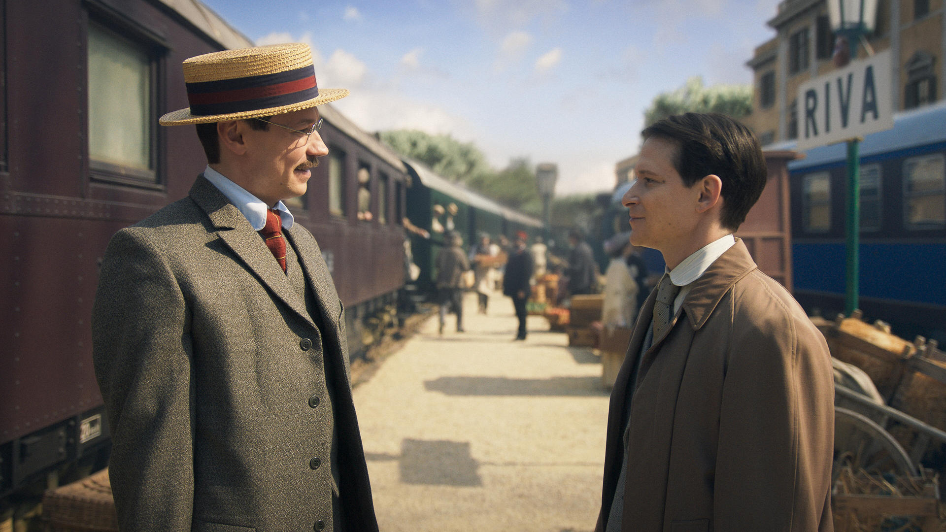 Zwei Männer in Vintage-Kleidung stehen sich auf einem Bahnsteig gegenüber. Ein Zug und ein Riva-Schild sind im Hintergrund zu sehen, zusammen mit anderen Personen.