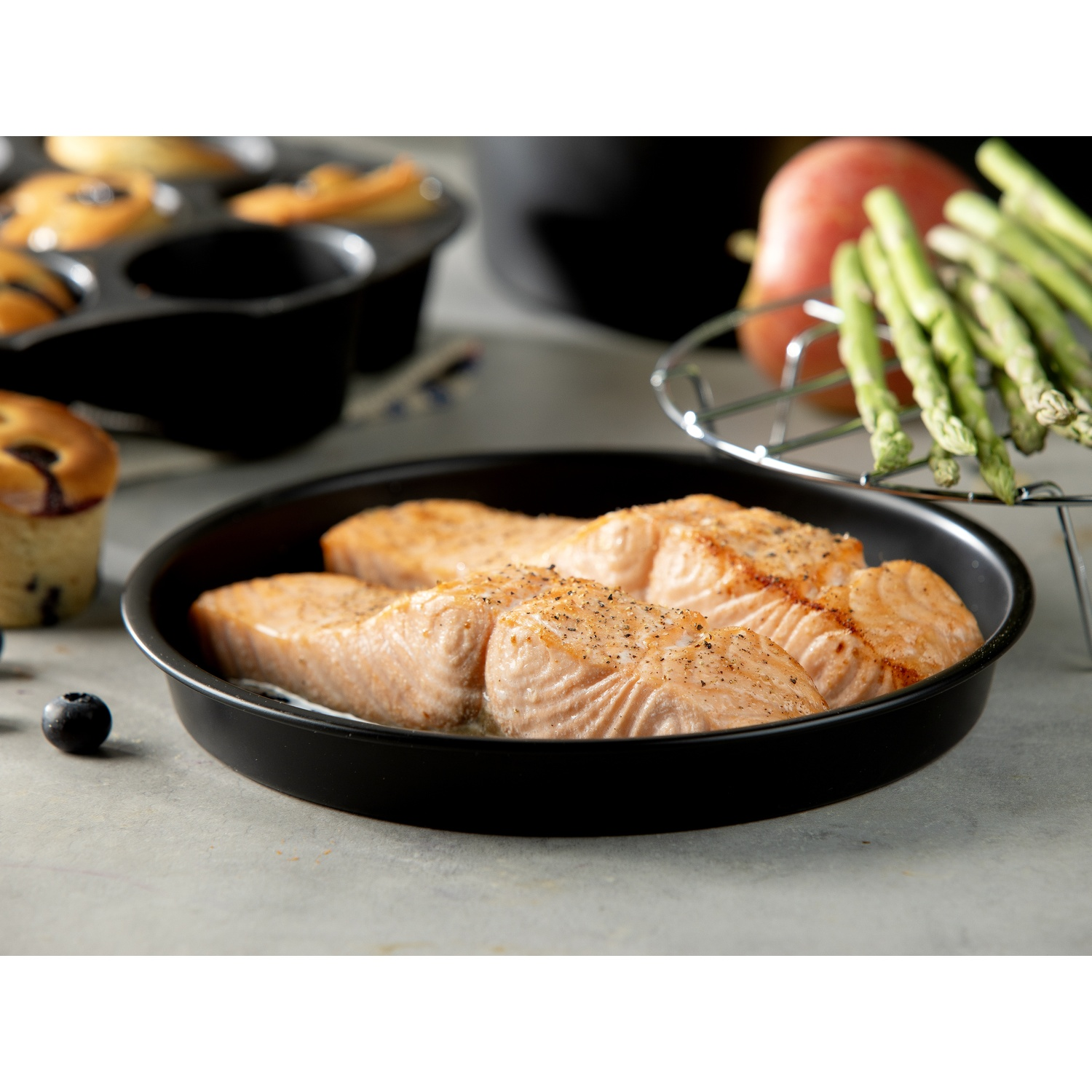 Pan-seared salmon in a black dish. Asparagus, pear, and muffins in the background. Blueberries scattered on a gray surface.