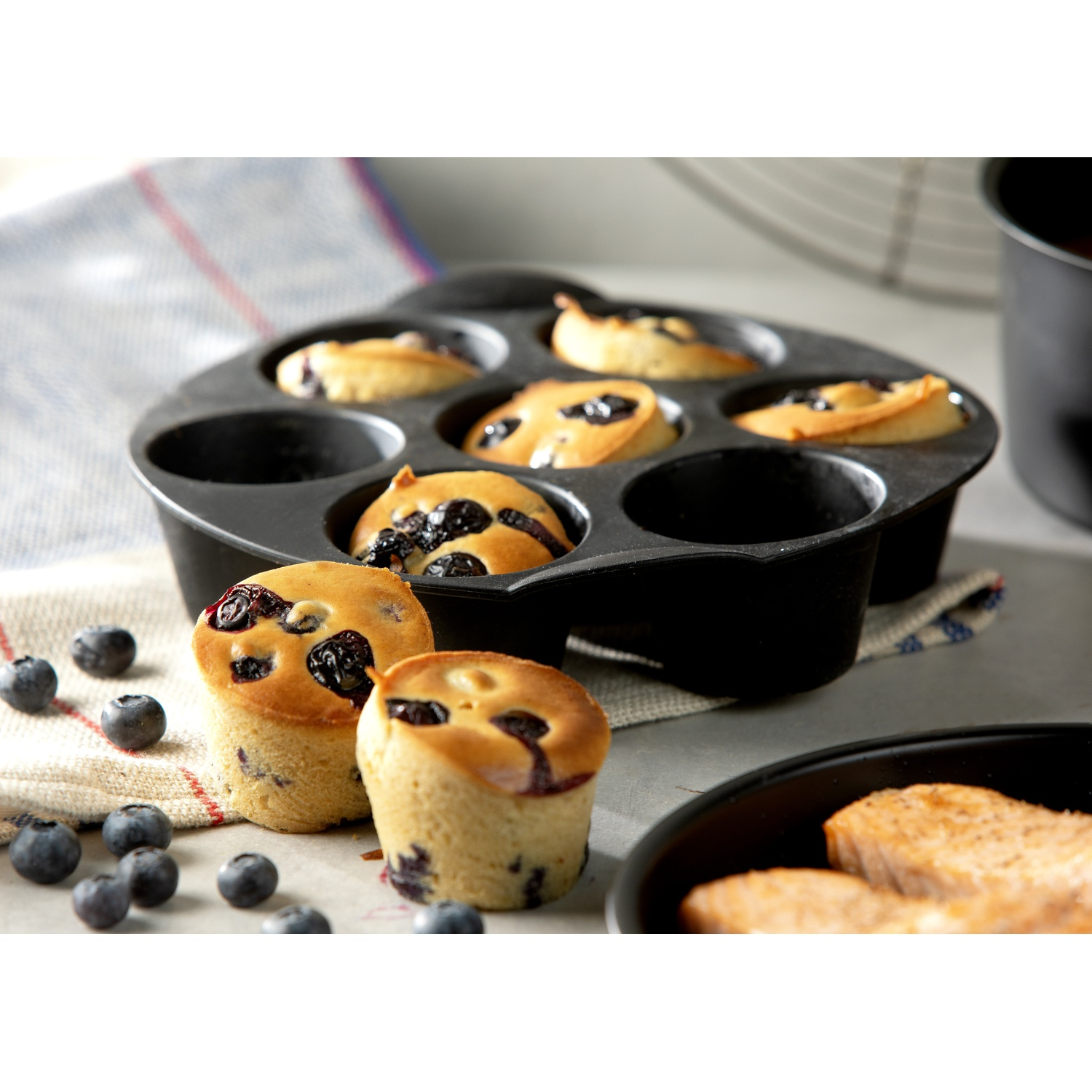 A muffin pan with baked blueberry muffins, fresh blueberries, and a plate of cooked salmon. A towel is in the background.