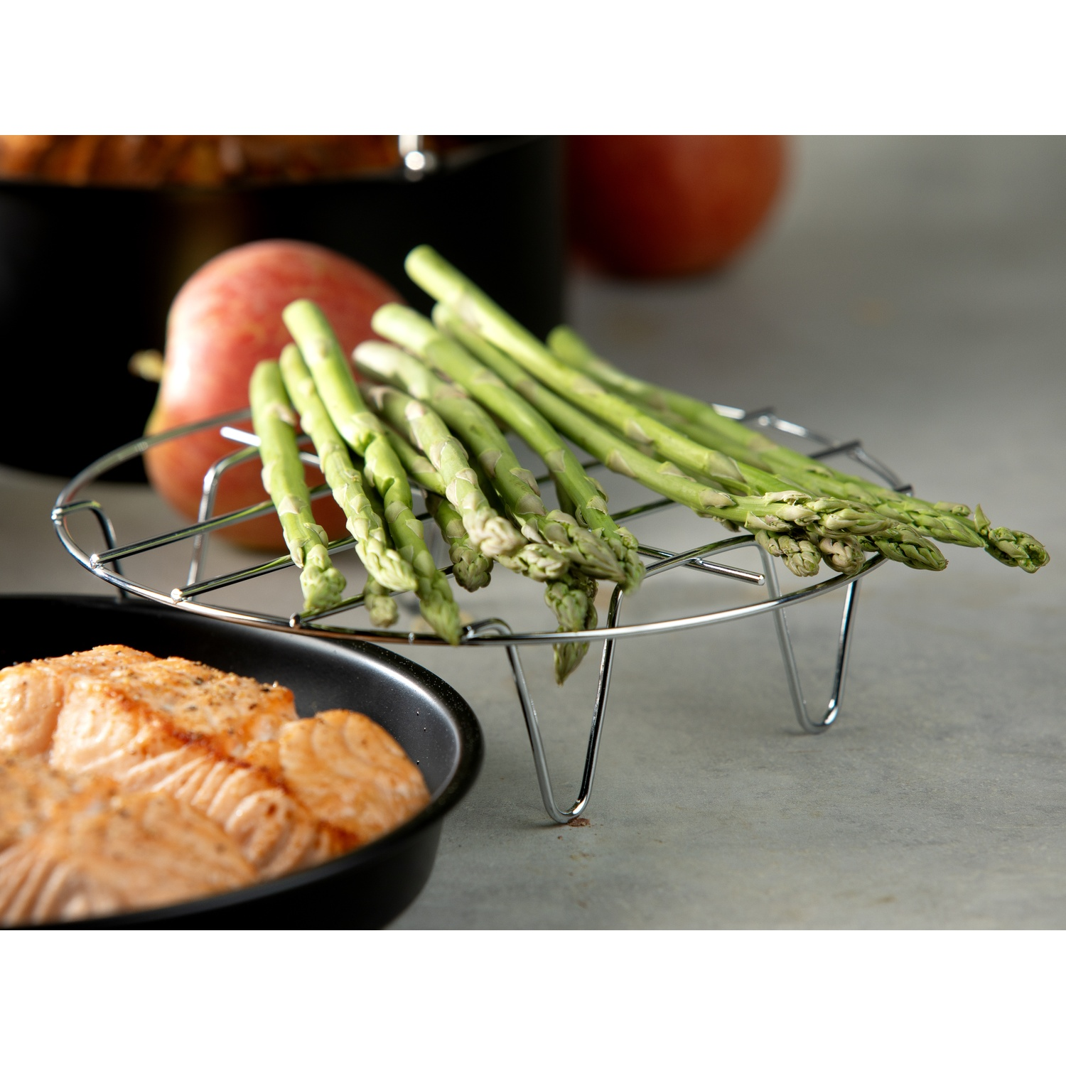 Asparagus on a wire rack, salmon in a pan, and an apple. The scene is set on a gray surface, suggesting a cooking context.