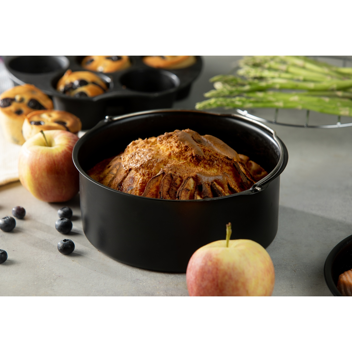A baked dish in a black pot, apples, blueberries, and muffins are on a gray surface with asparagus in the background.