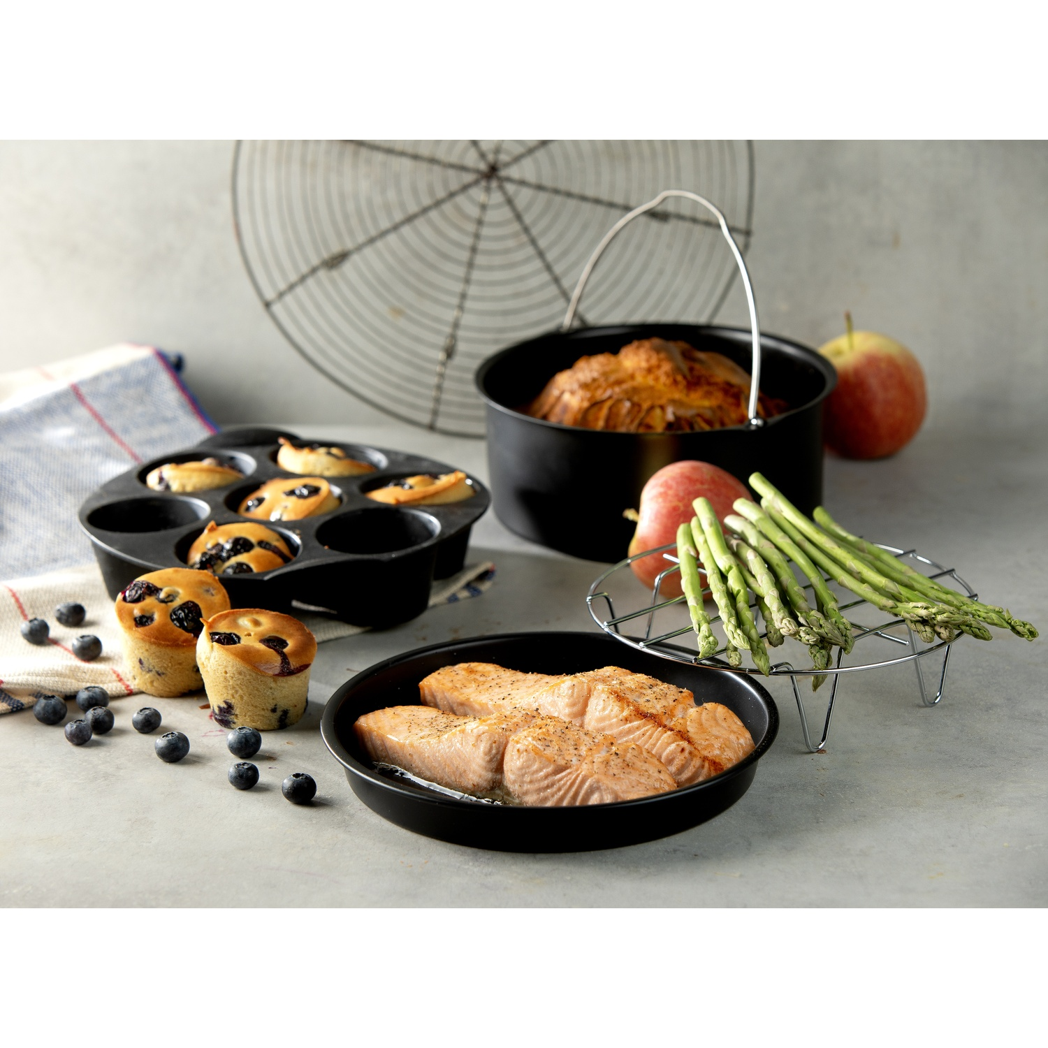 A table with muffins, salmon, apples, asparagus, and a baked dish. Black pans and a wire rack are also present.