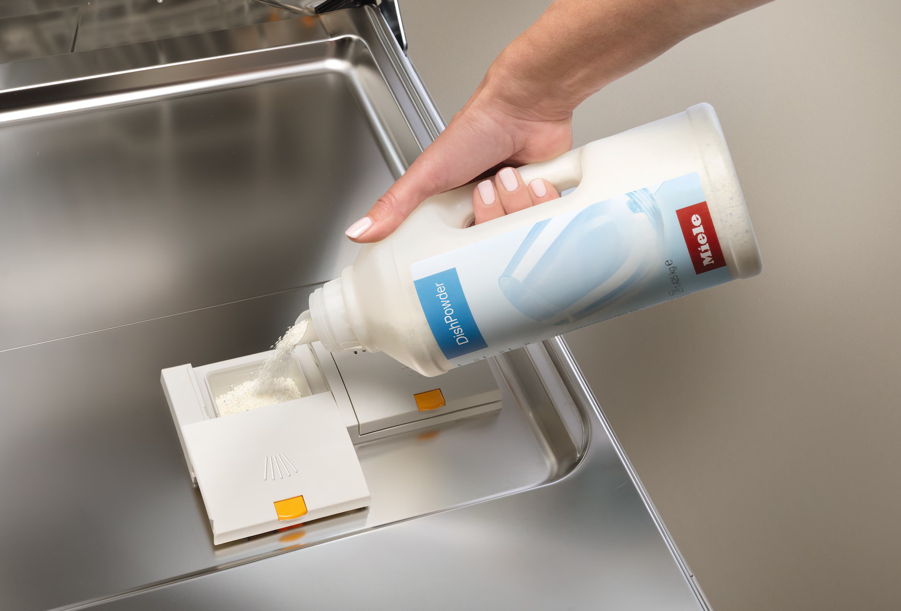 Eine Hand gießt Spülmittelpulver aus einer Miele-Flasche in einen Geschirrspülmittelspender. Der Spender ist weiß mit einem orangefarbenen Knopf.