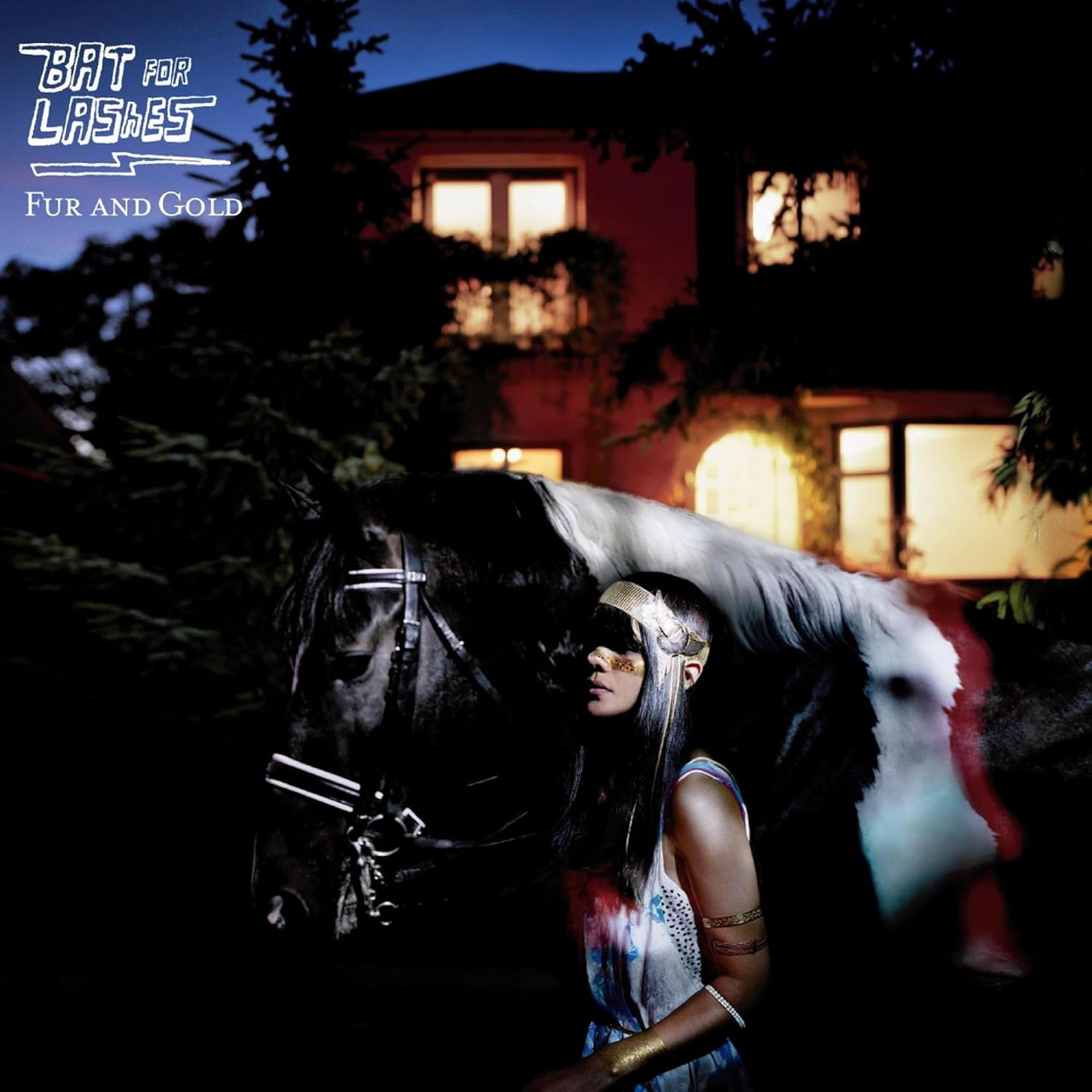 Nő arany fejdísszel és lóval. 'Bat for Lashes Fur and Gold' felirat. Ház a háttérben, világító ablakok, sötét ég.