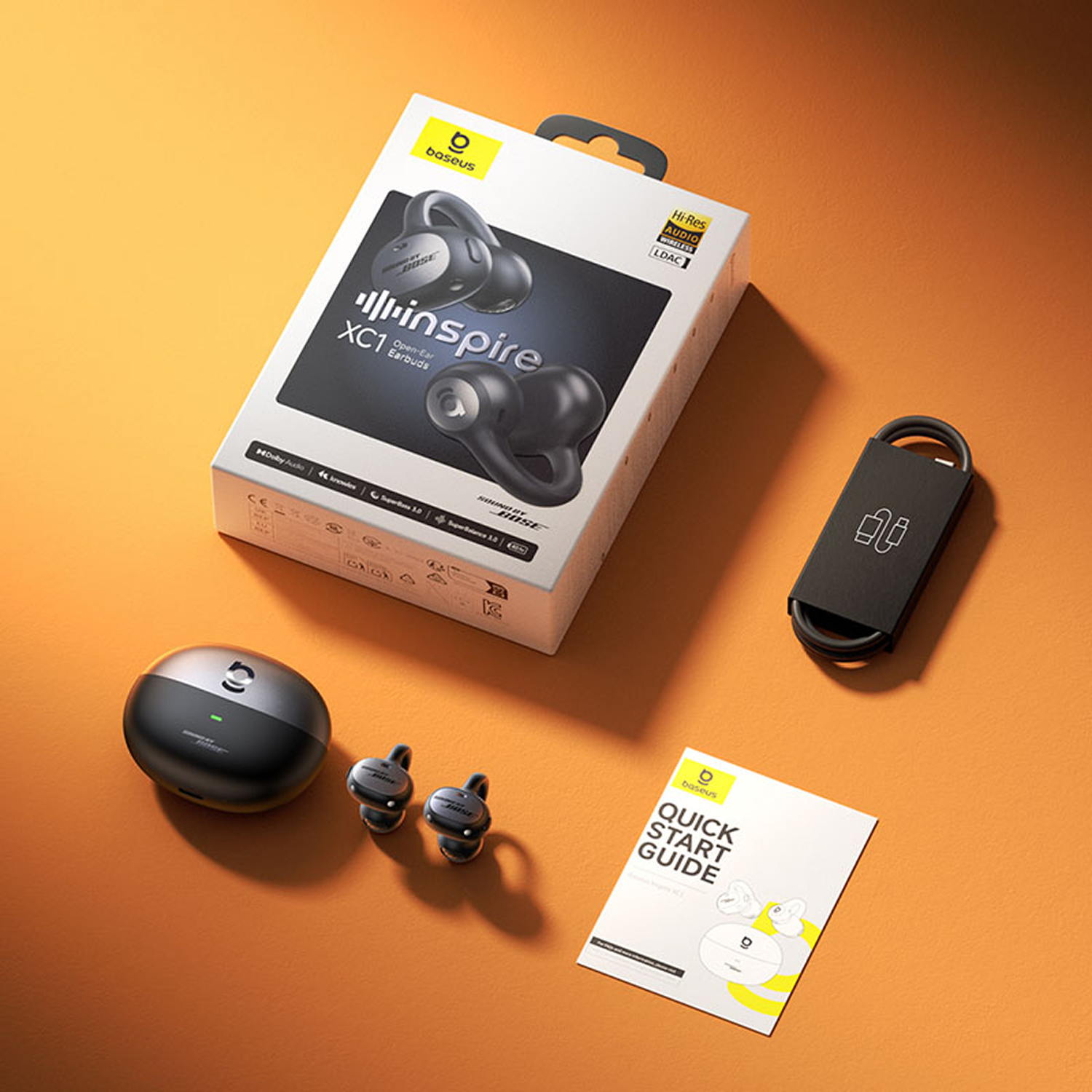 Caja de auriculares, cargador, auriculares y guía sobre superficie naranja. La caja dice 'Baseus Inspire XC1 Open-Ear Earbuds'. Hay auriculares negros.