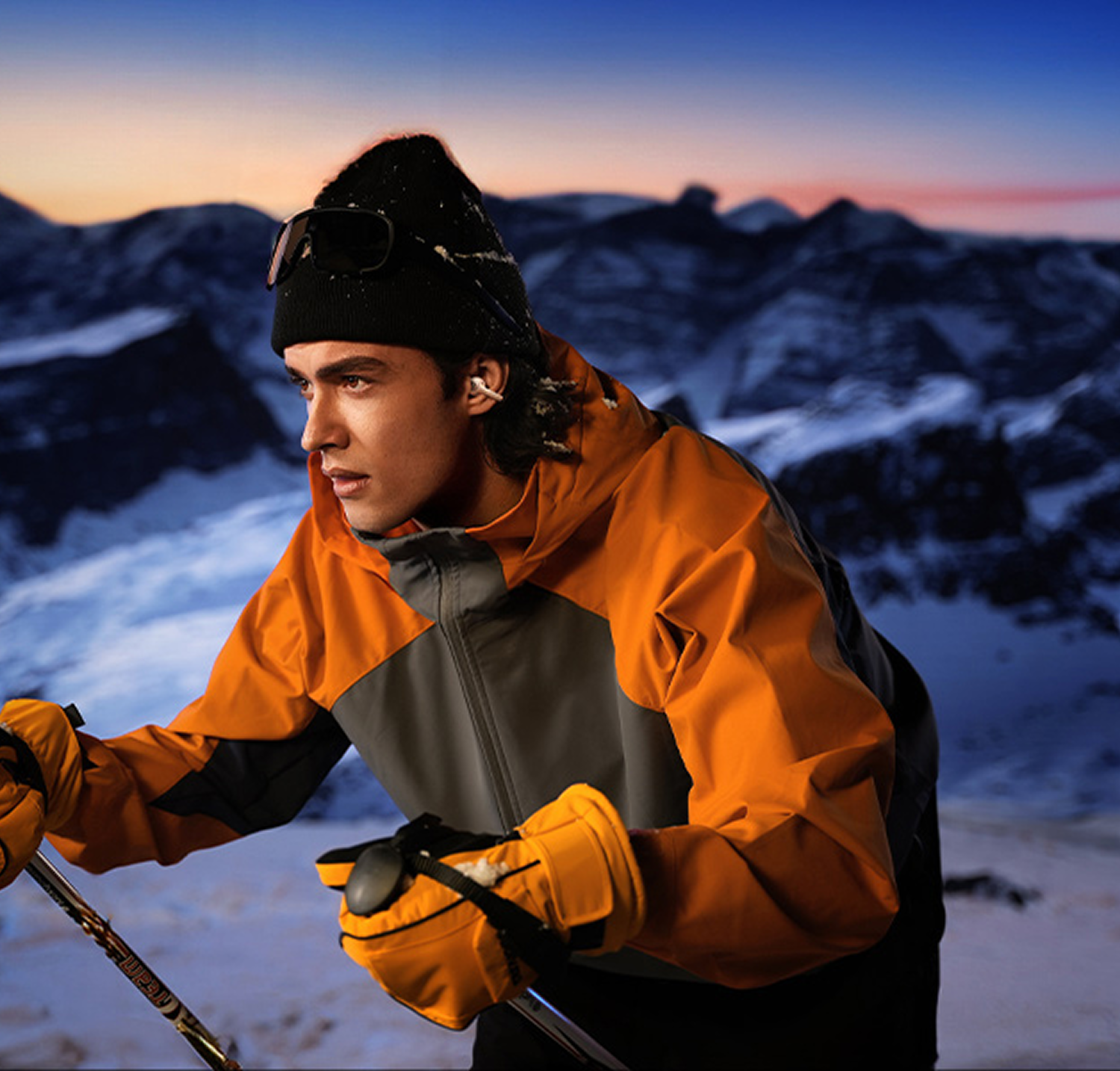 Esquiador con chaqueta naranja, gorro negro y guantes amarillos, sosteniendo bastones de esquí. Montañas nevadas al fondo, cielo al atardecer.