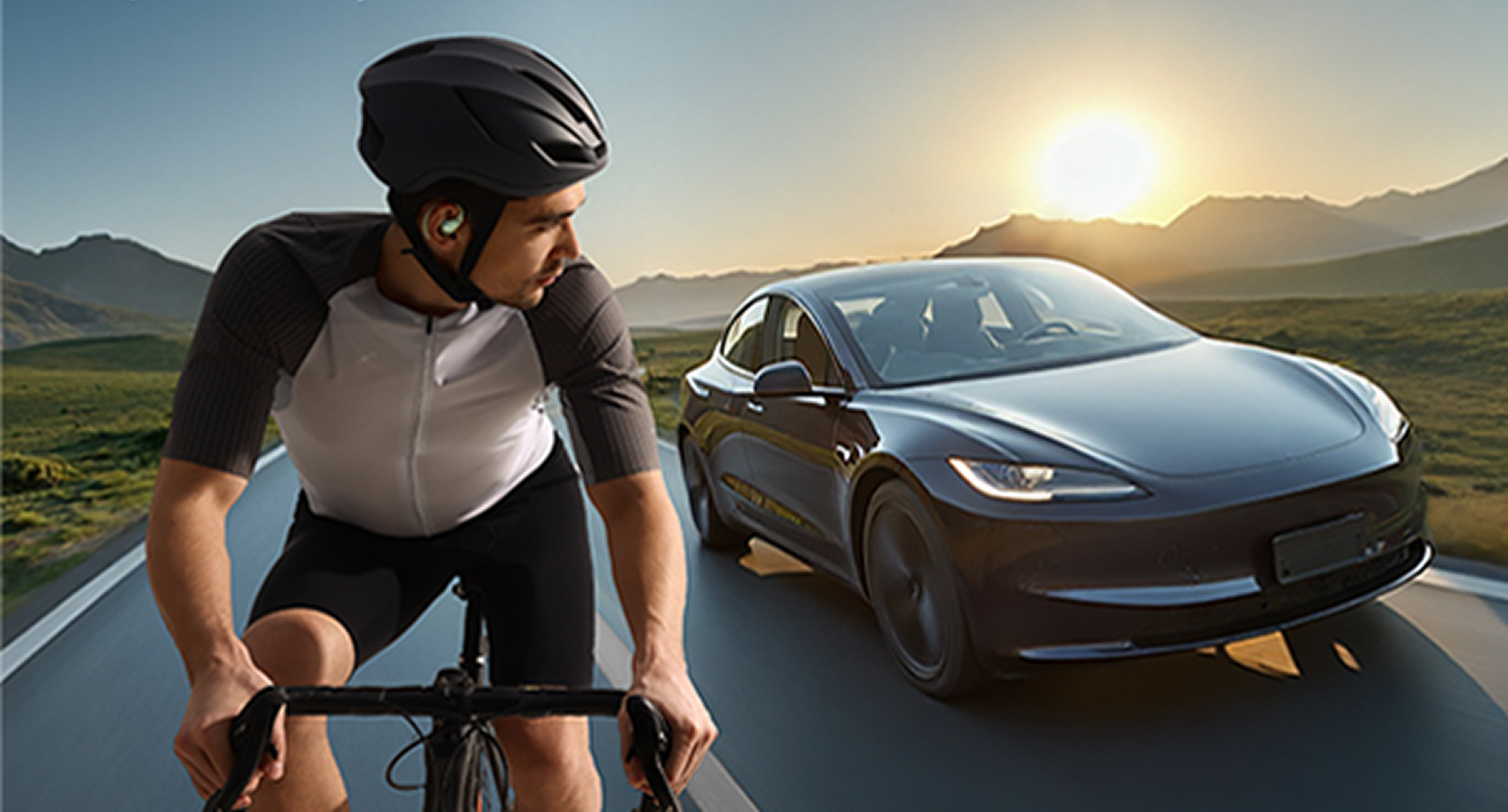 Ciclista con casco en bici, coche negro en carretera, montañas y sol al fondo.