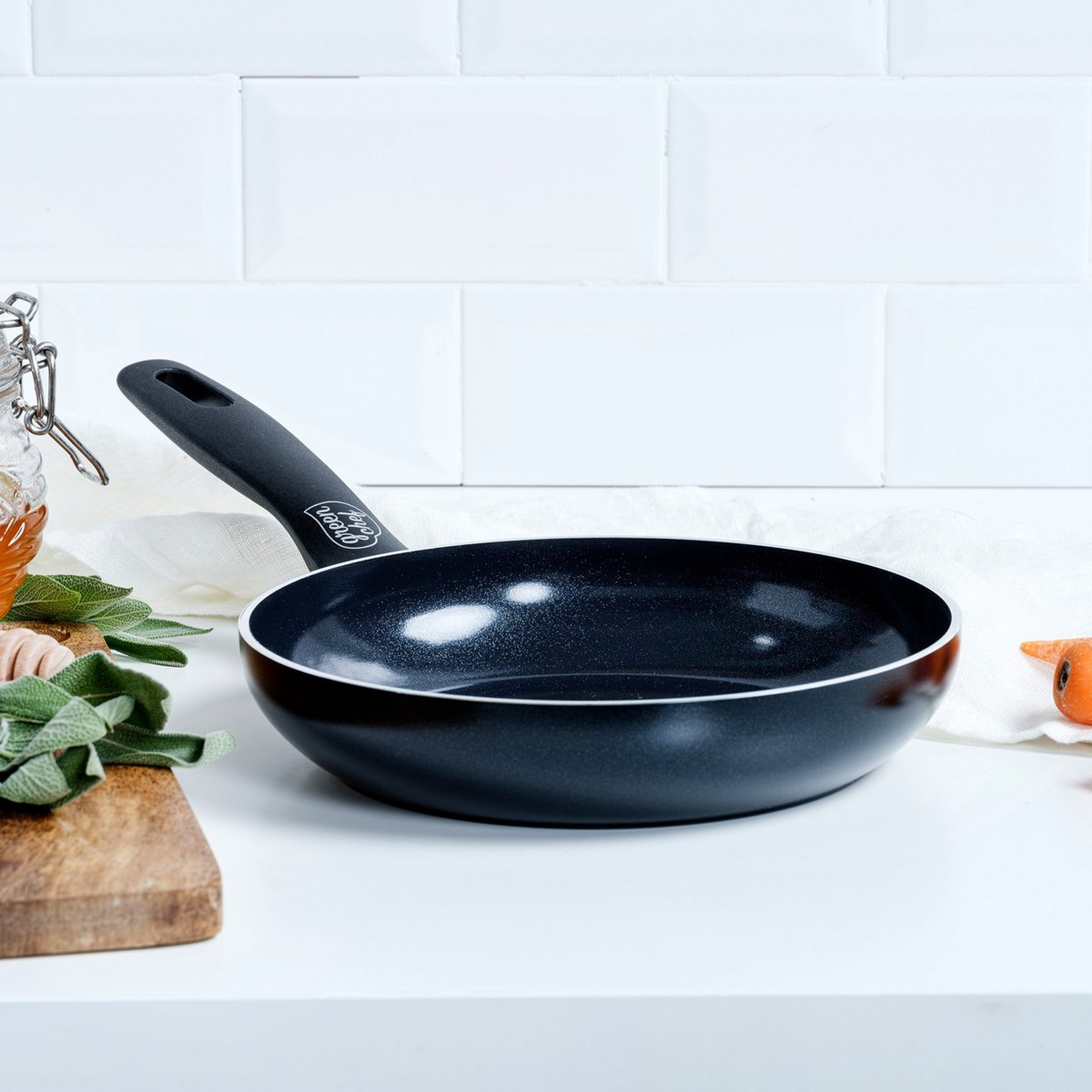 A black frying pan with a white rim sits on a white surface. Sage leaves, a wooden board, and a carrot are also visible.