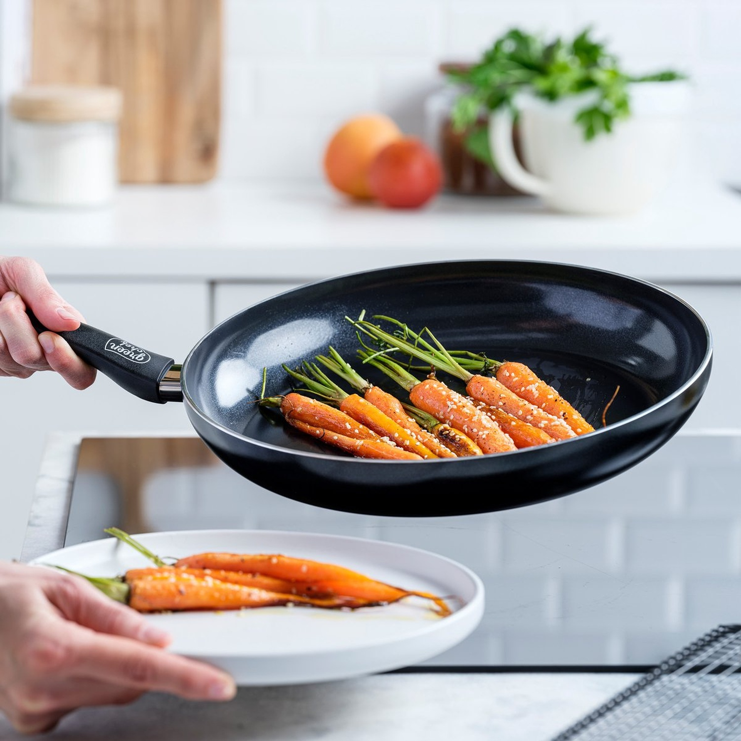 A person holding a pan of carrots with sesame seeds. A plate with carrots is below. Kitchen scene with fruits and herbs.