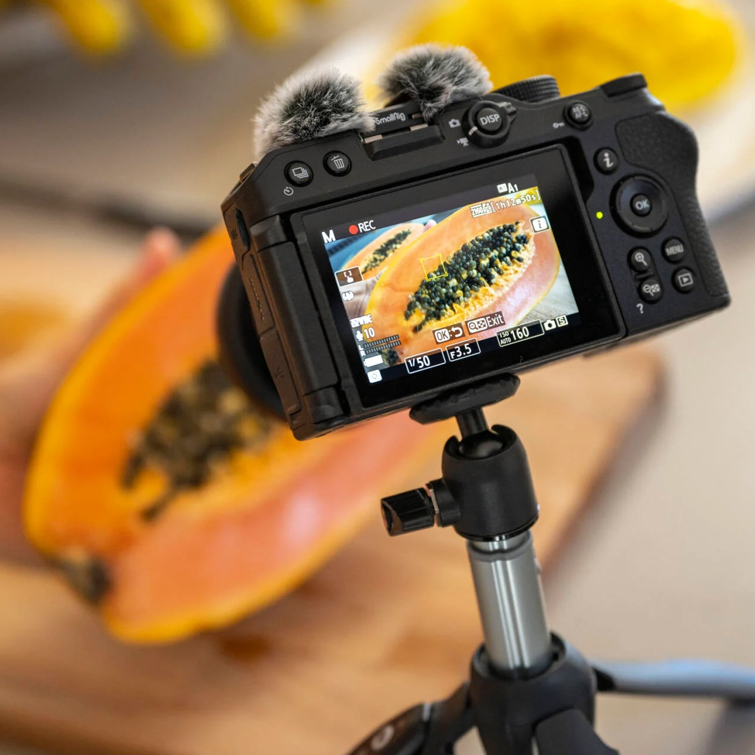 Una fotocamera su un treppiede mostra una papaya sul suo schermo. Una mezza papaya è sullo sfondo su una tavola di legno.