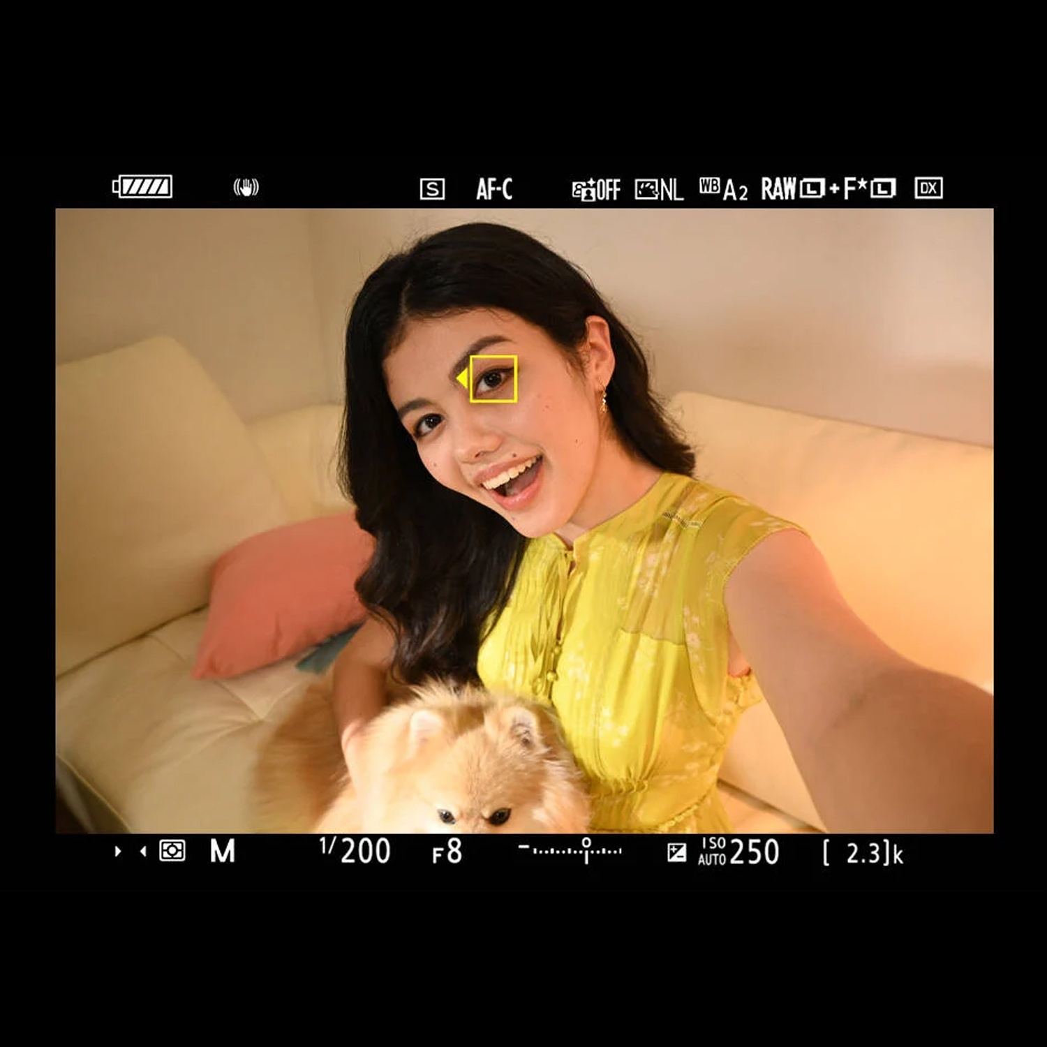 Una donna sorride, tenendo un cane soffice. Indossa il giallo, seduta su un divano. Le impostazioni della fotocamera sono visibili.