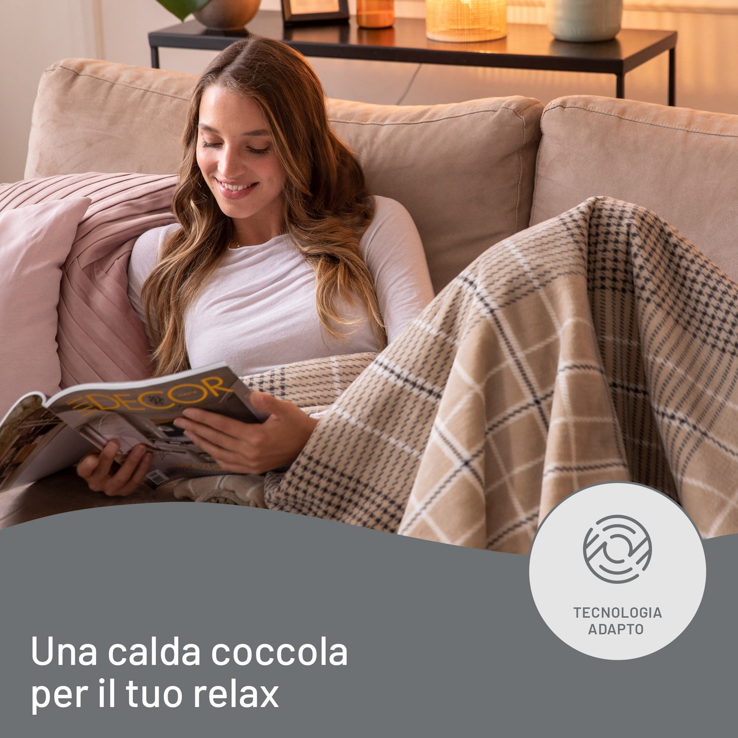 Donna su un divano che legge una rivista, coperta da una coperta. C'è un cuscino decorativo. Lo sfondo mostra un tavolo e una lampada.