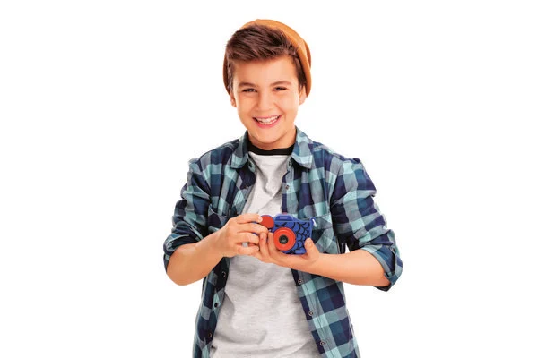 Un niño sonriente con una camisa a cuadros y gorro sostiene una cámara de juguete. Está frente a un fondo blanco.