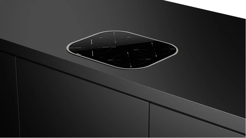 Una placa de inducción negra con cinco quemadores está instalada en una encimera oscura. La cocina es moderna y minimalista.