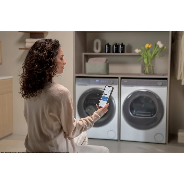 Una mujer en el lavadero mira su teléfono para controlar electrodomésticos. Dos lavadoras/secadoras al fondo, con flores y suministros.