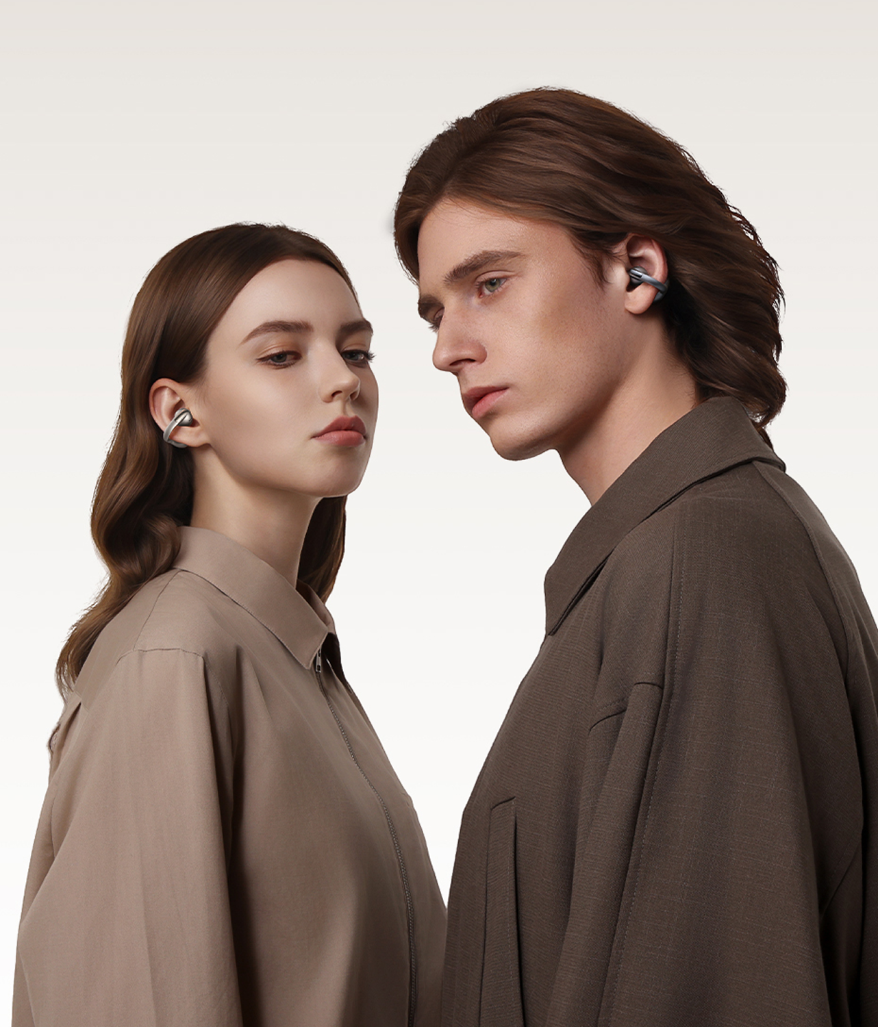 Una mujer y un hombre con auriculares, vestidos con camisas de tonos neutros, mirándose sobre un fondo blanco.