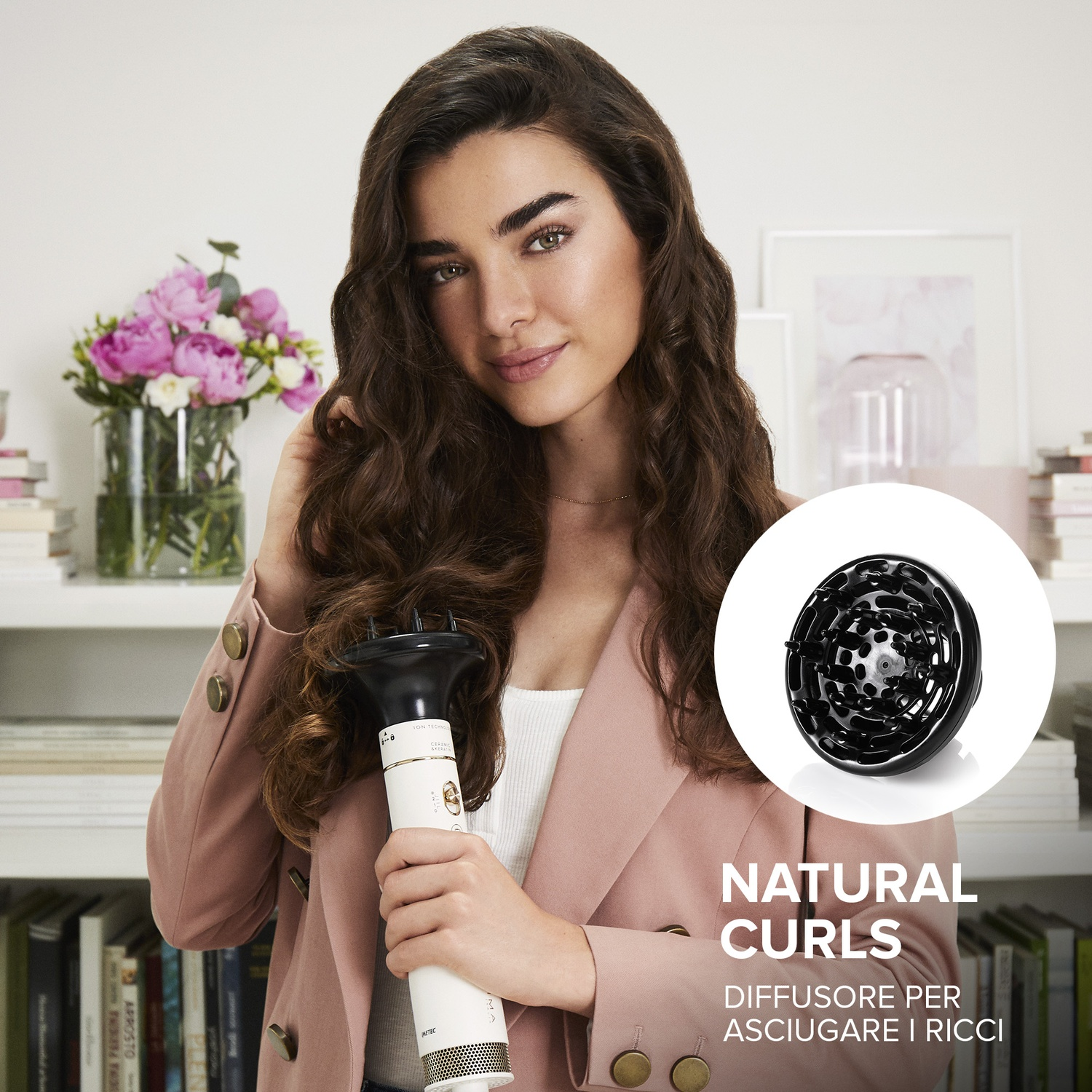 Donna con capelli ricci tiene un diffusore per capelli. Un'arrangiamento floreale e libri sullo sfondo.