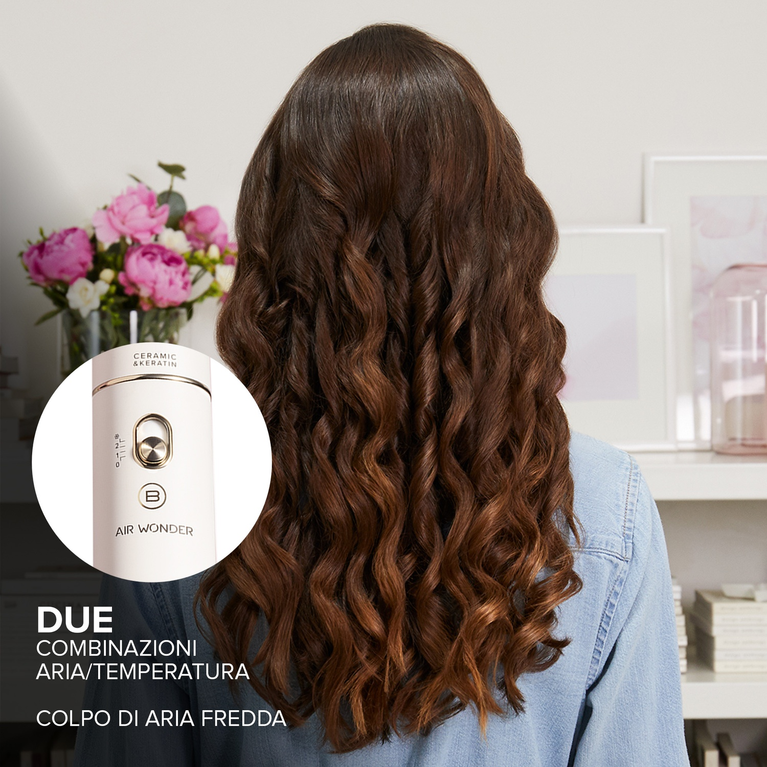 Donna con lunghi capelli castano ondulati, viene mostrato uno strumento per lo styling dei capelli. Fiori rosa in un vaso su uno scaffale dietro di lei.