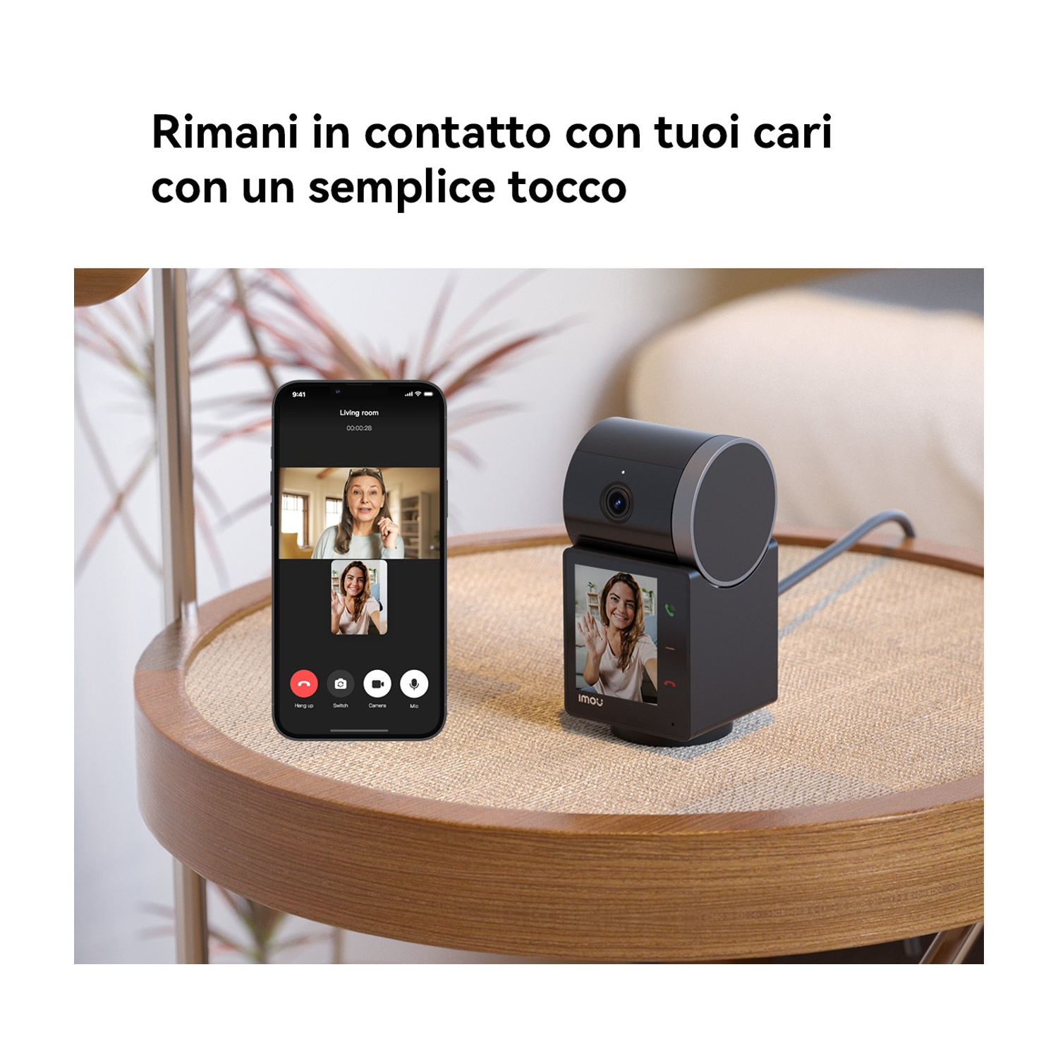 Un telefono e una telecamera su un tavolo mostrano una videochiamata. Una donna sulla telecamera saluta. Il testo in alto recita: 'Rimani in contatto con i tuoi cari'.